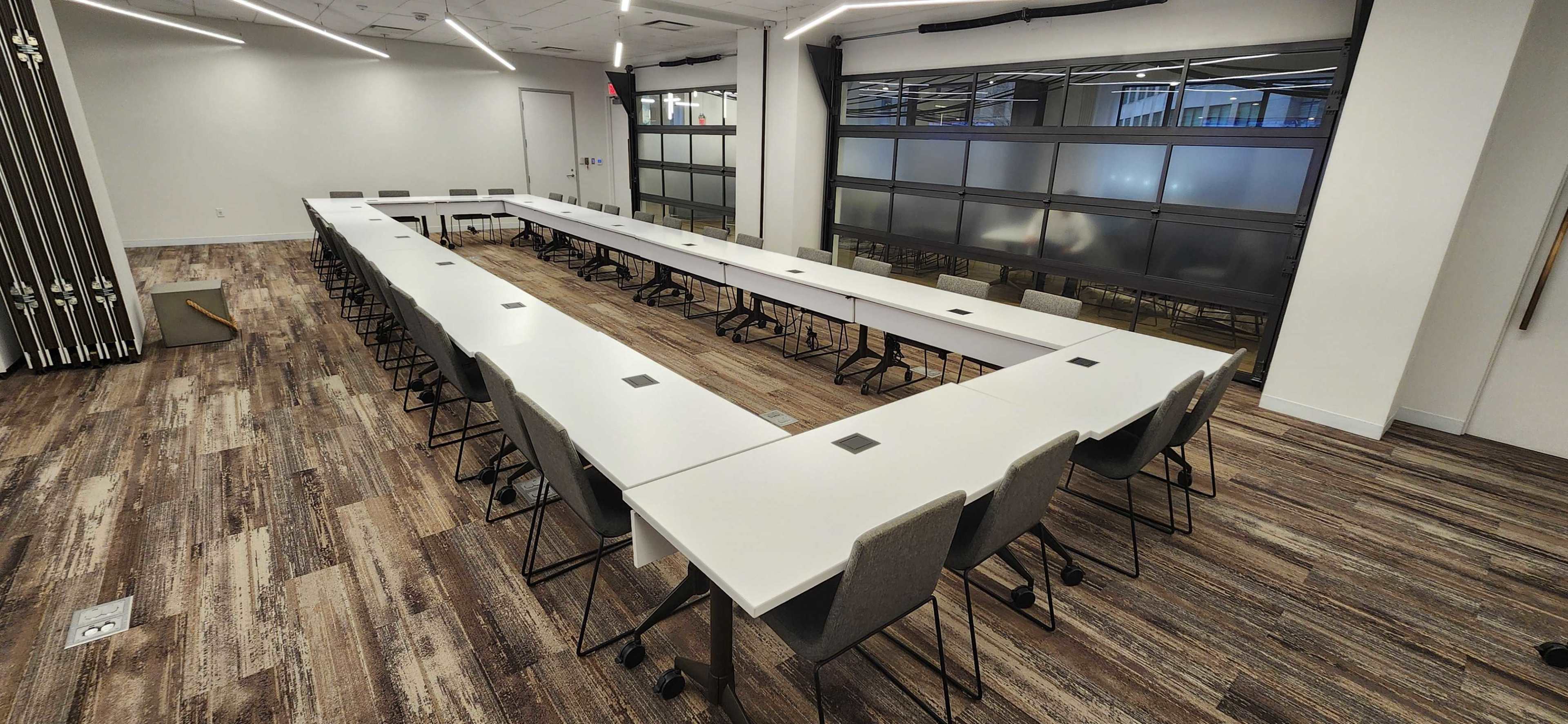 A conference room features a large U-shaped table arrangement with chairs and a wooden floor under fluorescent lighting.