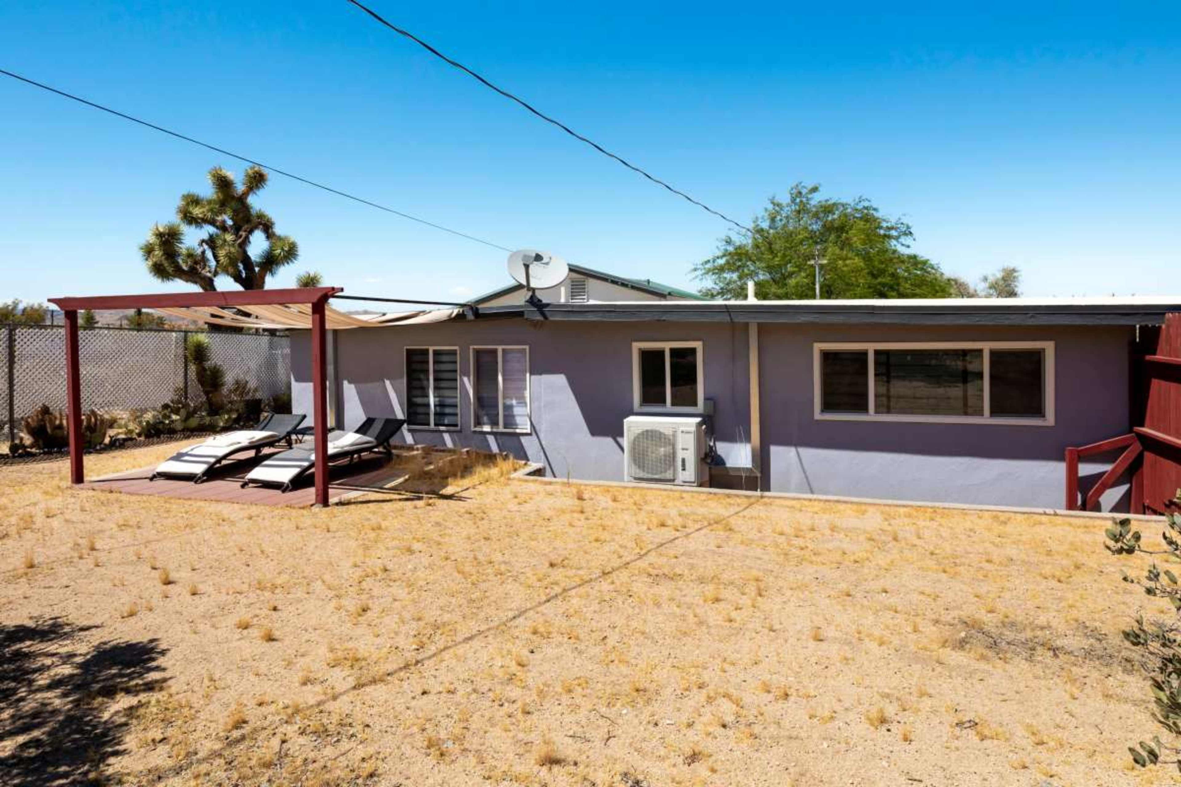 A simple, single-story house with a covered patio and two lounge chairs stands in a sandy, barren yard under a clear blue sky.