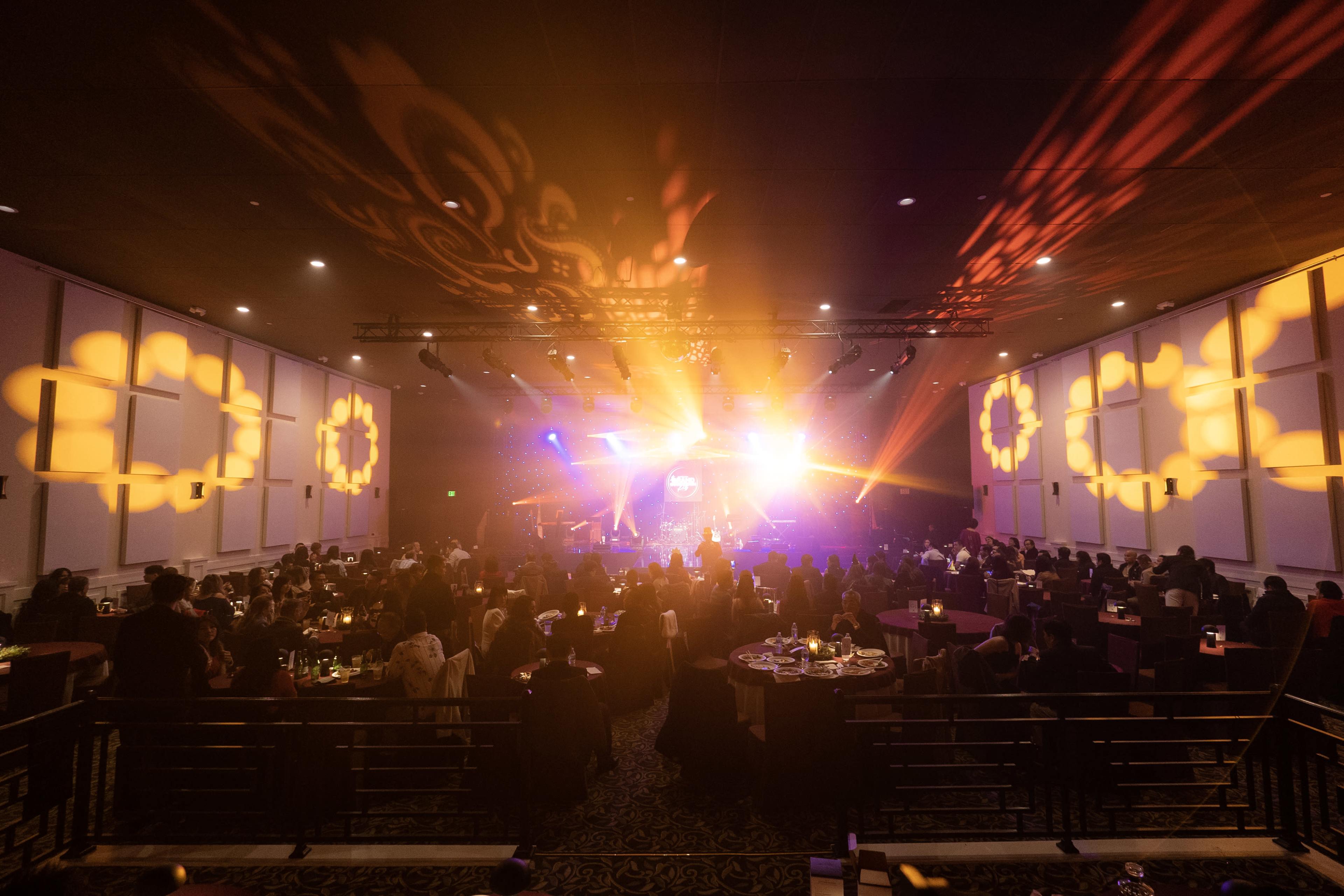 A crowd gathers in a dimly lit event space with vibrant lights and projections illuminating the walls and stage.