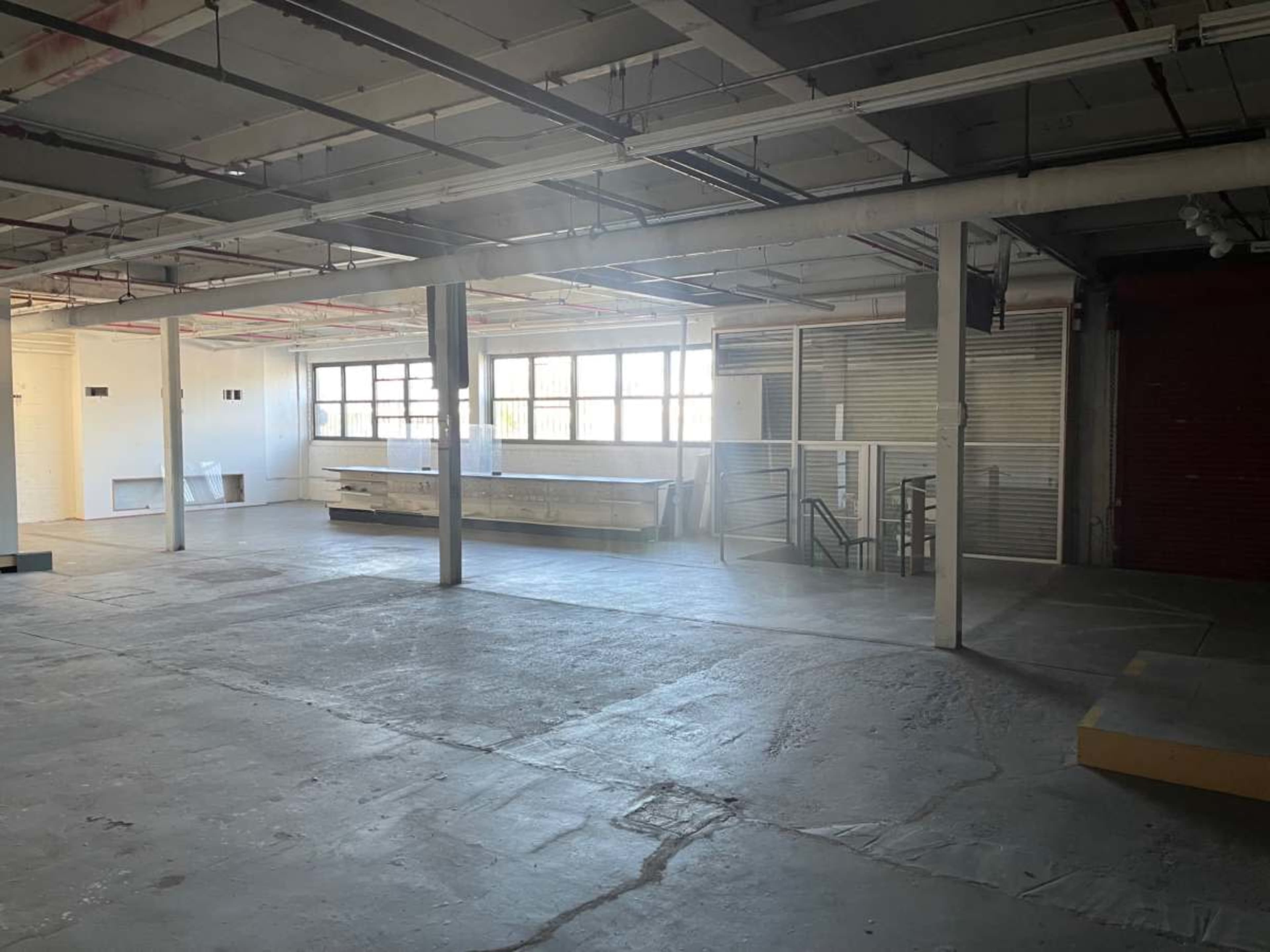 The image shows a spacious, empty industrial room with large windows and exposed beams.