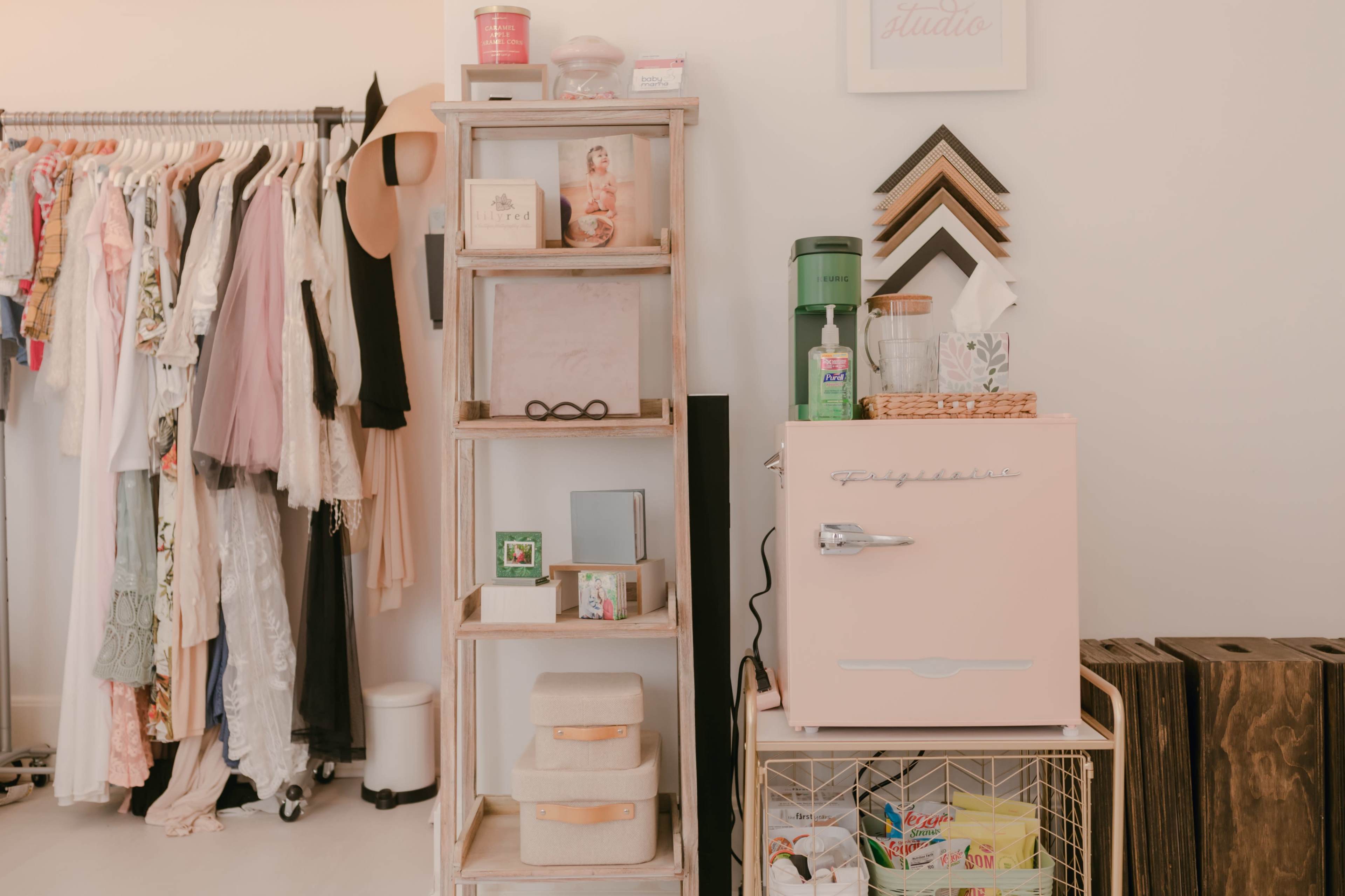 The image shows a cozy room with a clothing rack on the left, a wooden shelf displaying various items in the center, and a pink refrigerator on the right.