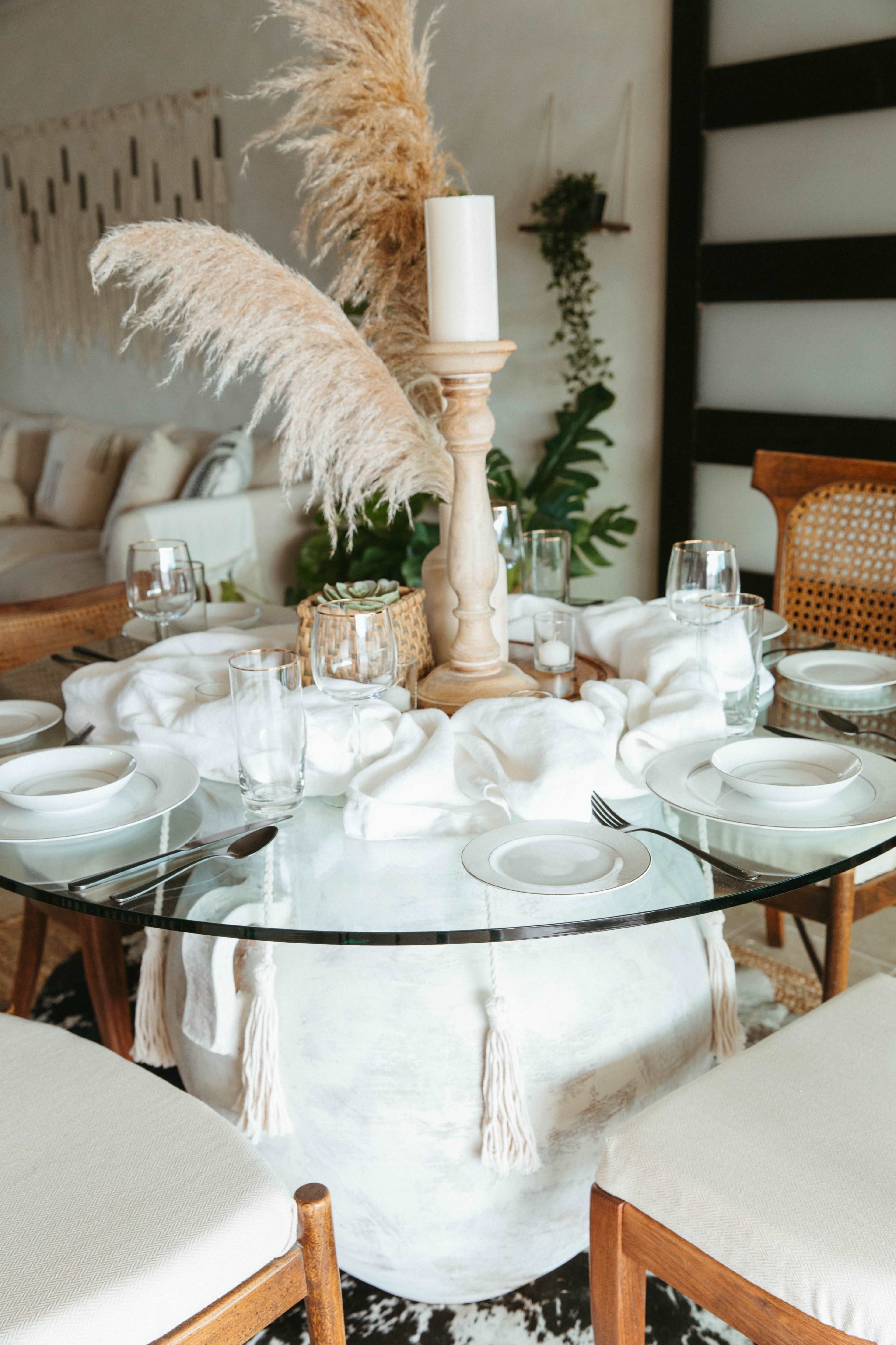 A round dining table is set with white plates and glasses, featuring a tall wooden candle holder and decorative pampas grass in the center.