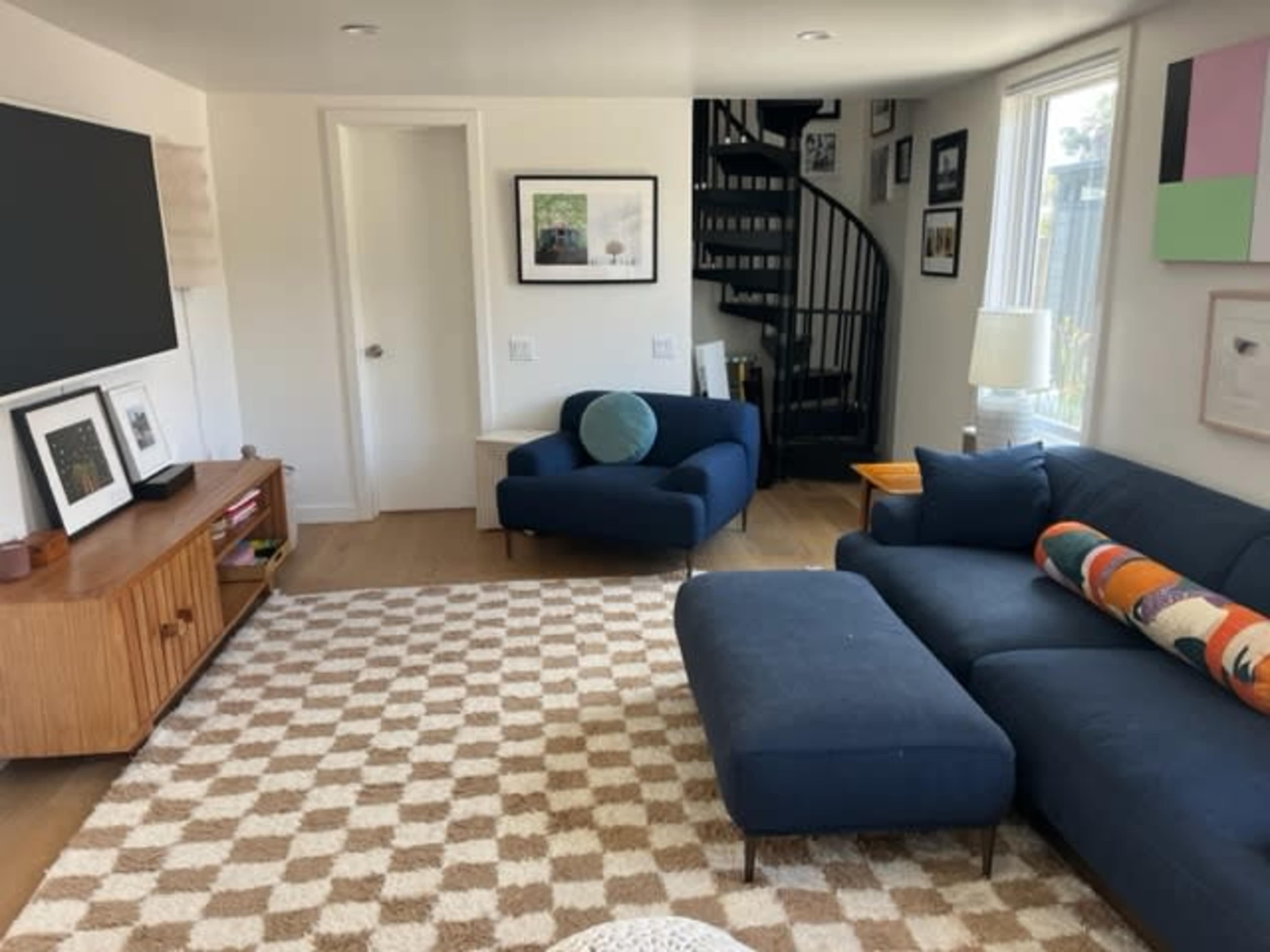 A modern living room with a sectional sofa, a blue armchair, a wooden TV cabinet, a patterned rug, and a spiral staircase.
