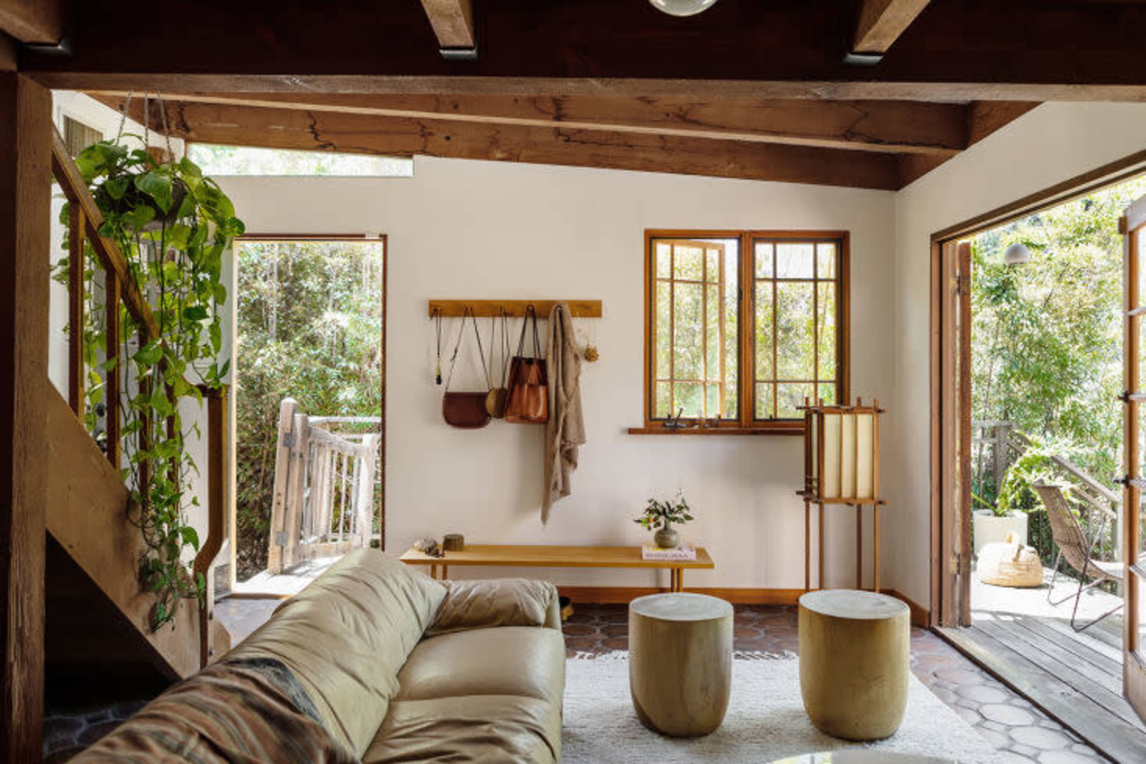The image depicts a cozy living room with wooden beams, a large sofa, two round tables, and large windows opening to a green outdoor area.