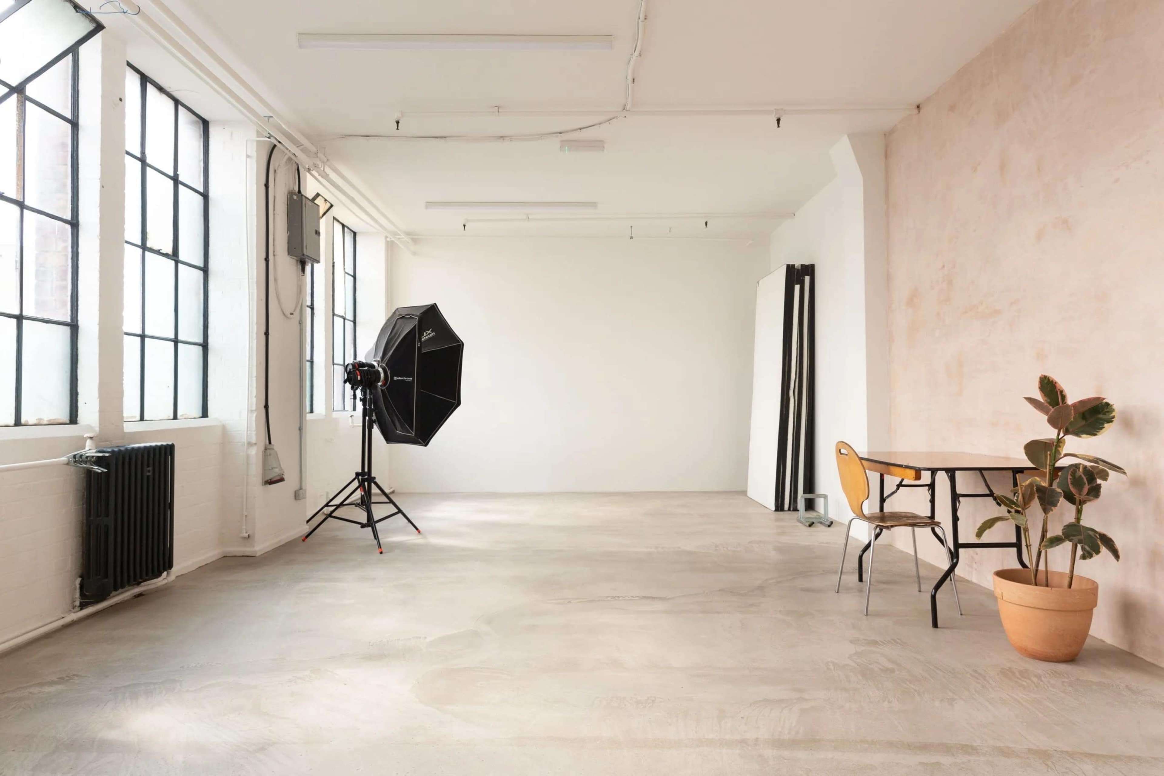 The image shows a spacious, minimalist studio with large windows, a lighting setup, a table, and a potted plant.