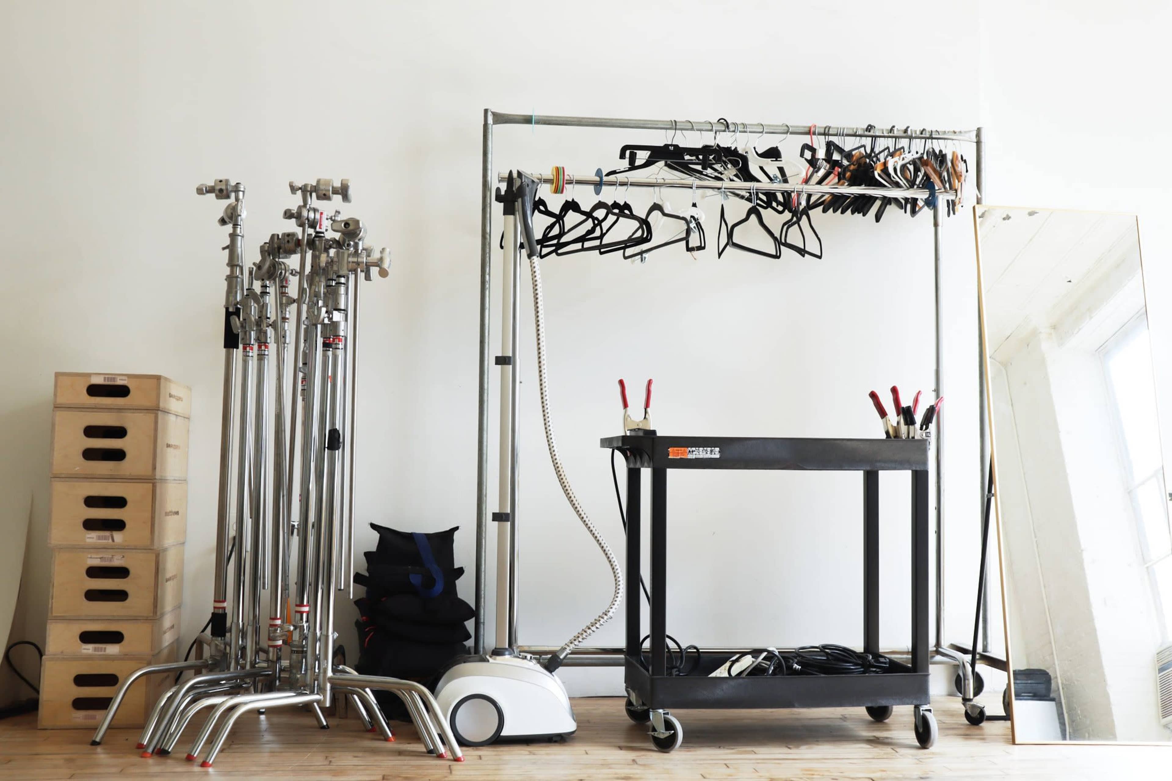 The image shows a clean, organized studio space with a clothing rack holding hangers, a metal cart with clamps, a steamer, and several tall stands at the side.