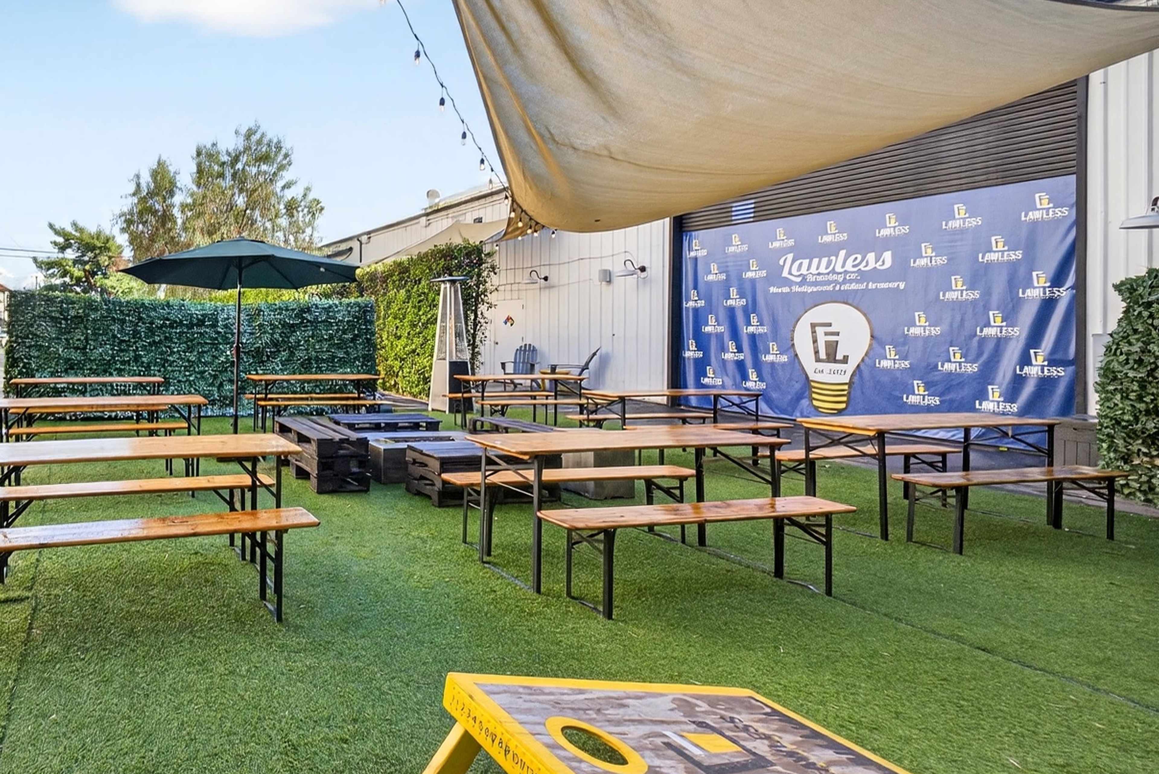 The image shows an outdoor event space with multiple wooden benches and tables arranged on green turf, alongside a wall featuring a branded backdrop for "Lawless."