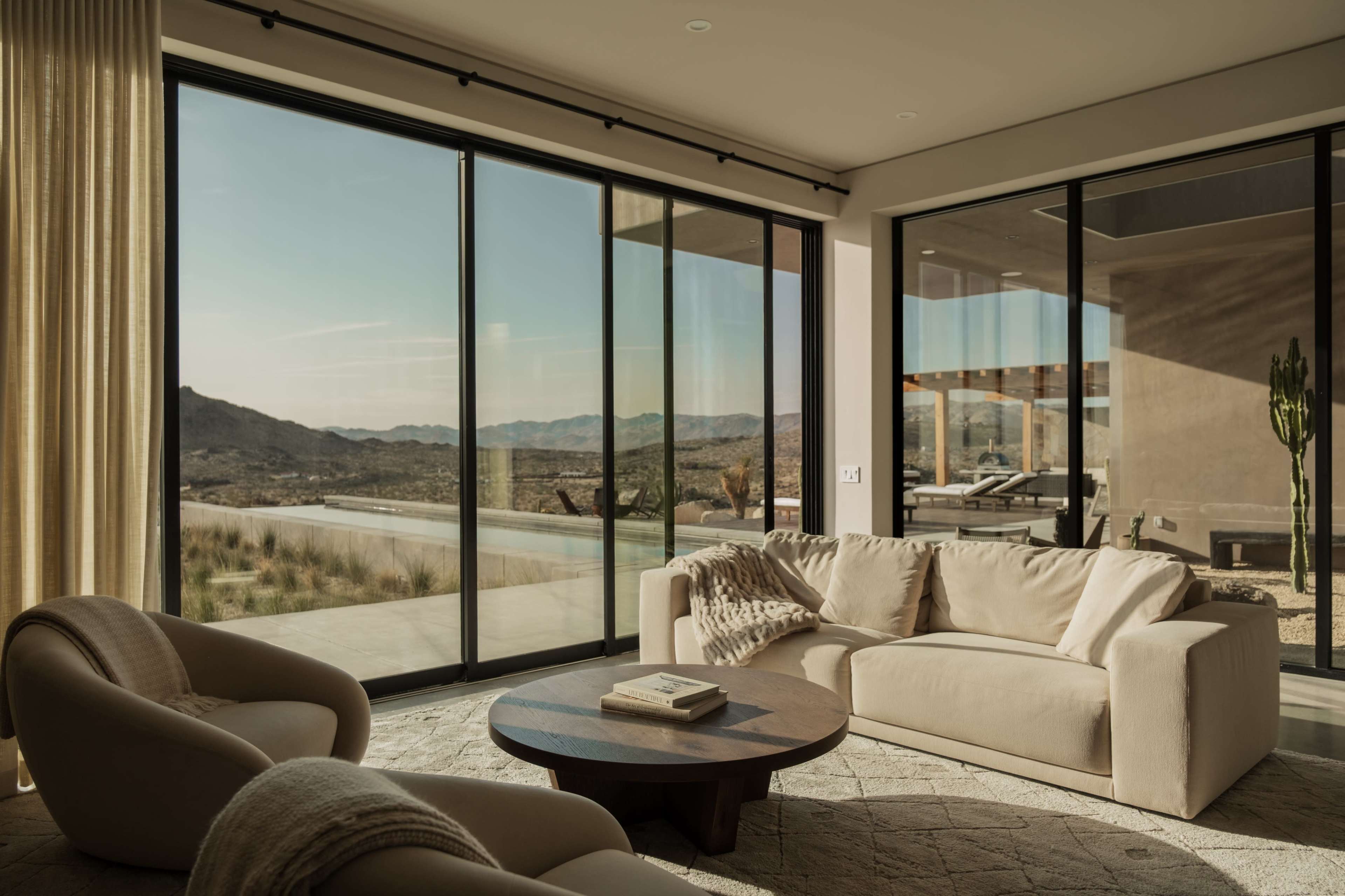 A modern living room features large glass windows overlooking a desert landscape with mountains in the distance.