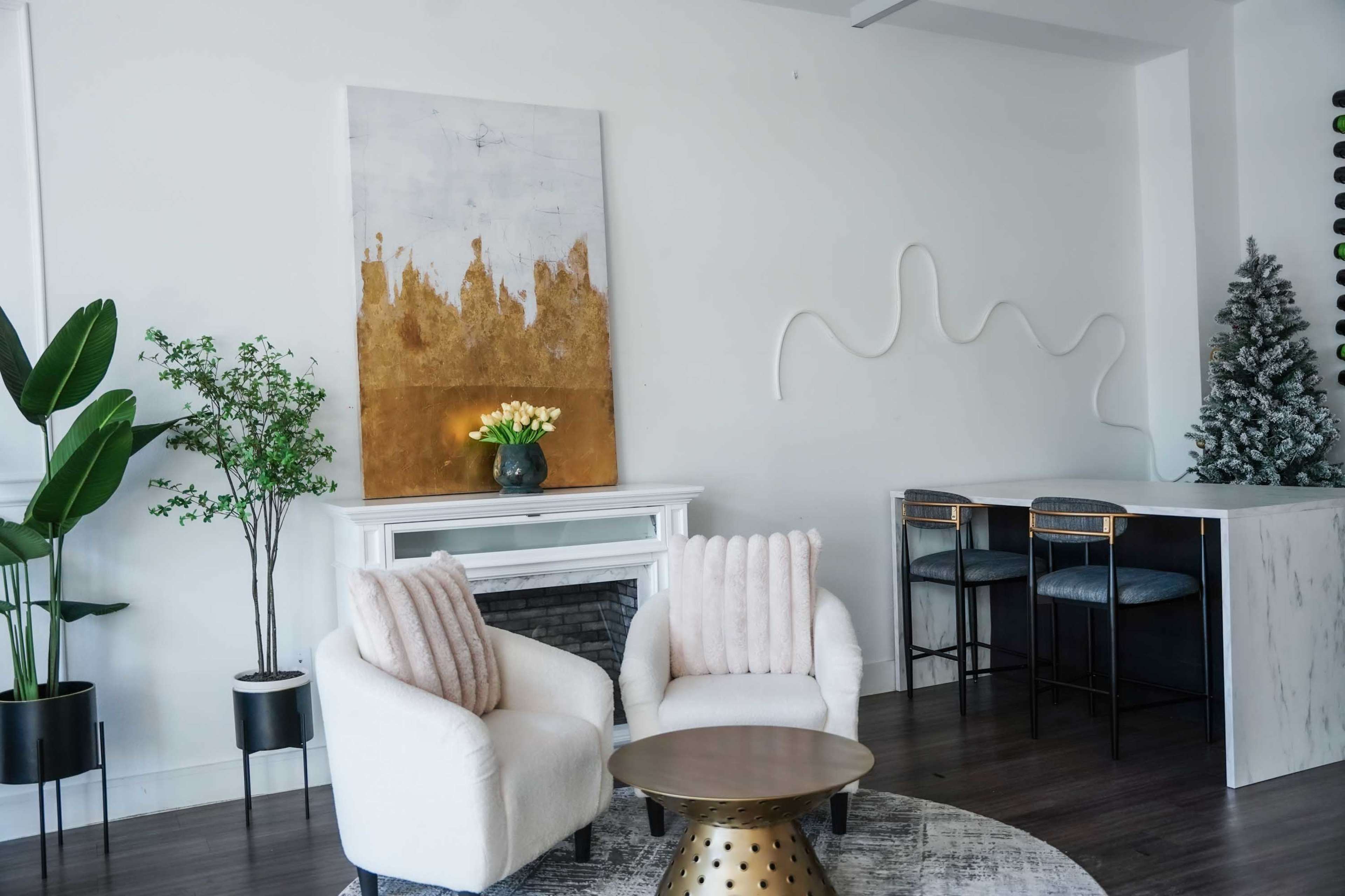 The image depicts a modern living space featuring two white armchairs, a round coffee table with a patterned design, and a wall decorated with a large abstract painting and a minimalist design element.
