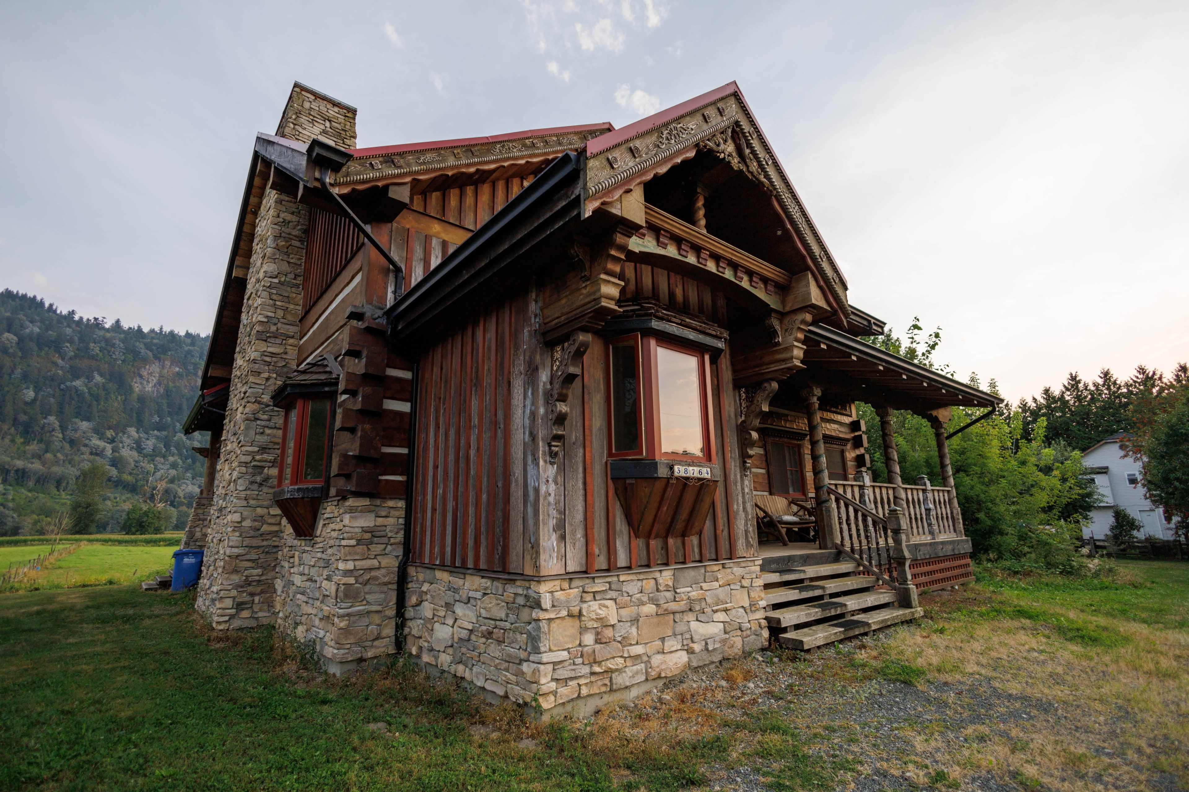 The image shows a rustic house made of wood and stone, featuring intricate architectural details and a prominent porch.