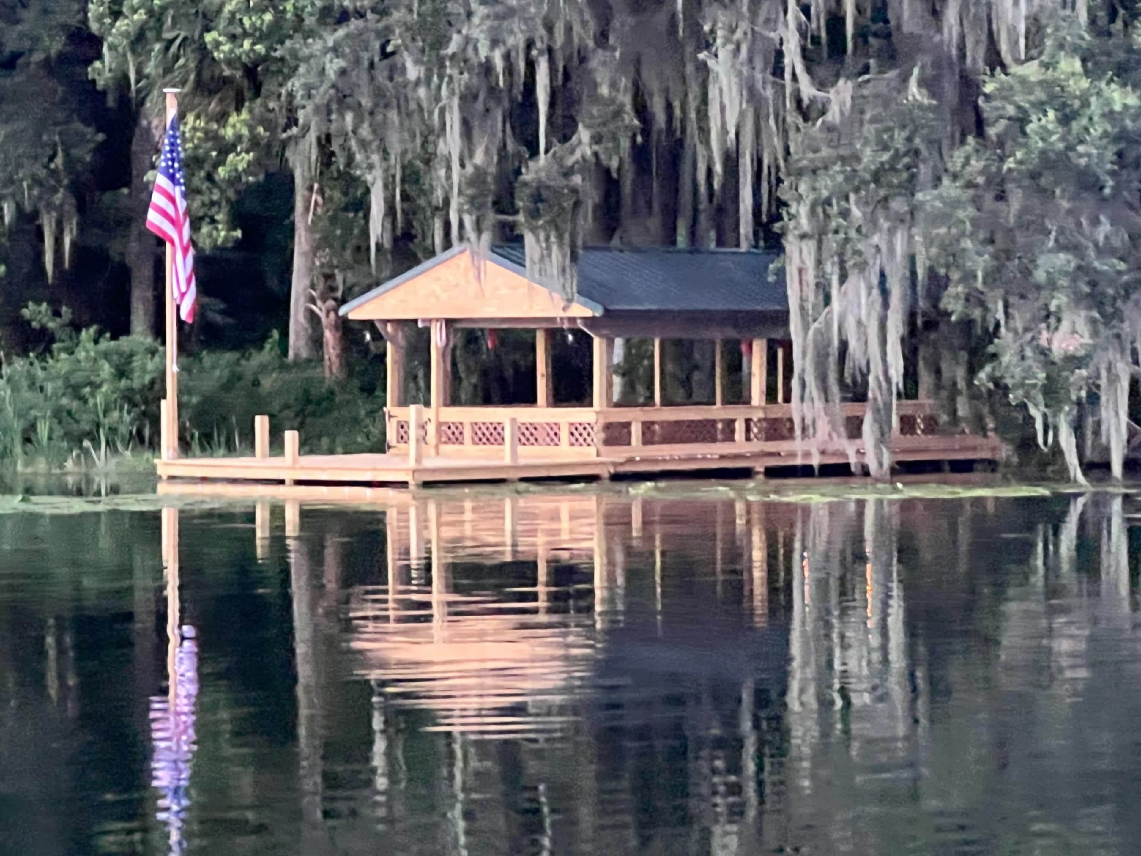 A wooden dock with a covered seating area is situated on a serene body of water, surrounded by trees draped in Spanish moss, with an American flag waving nearby.