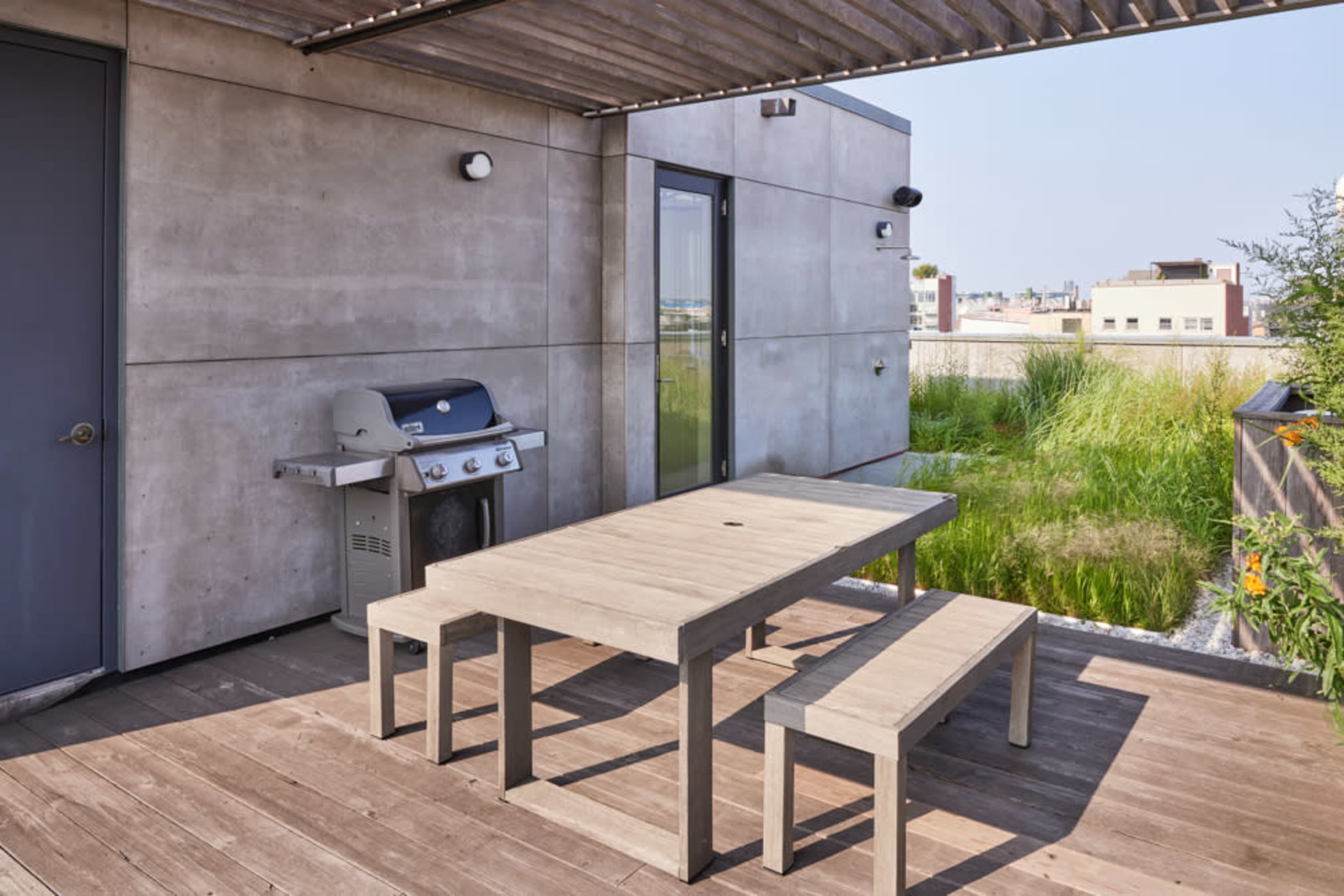 A rooftop terrace with a wooden table and benches beside a barbecue grill, surrounded by green plants and a clear sky.