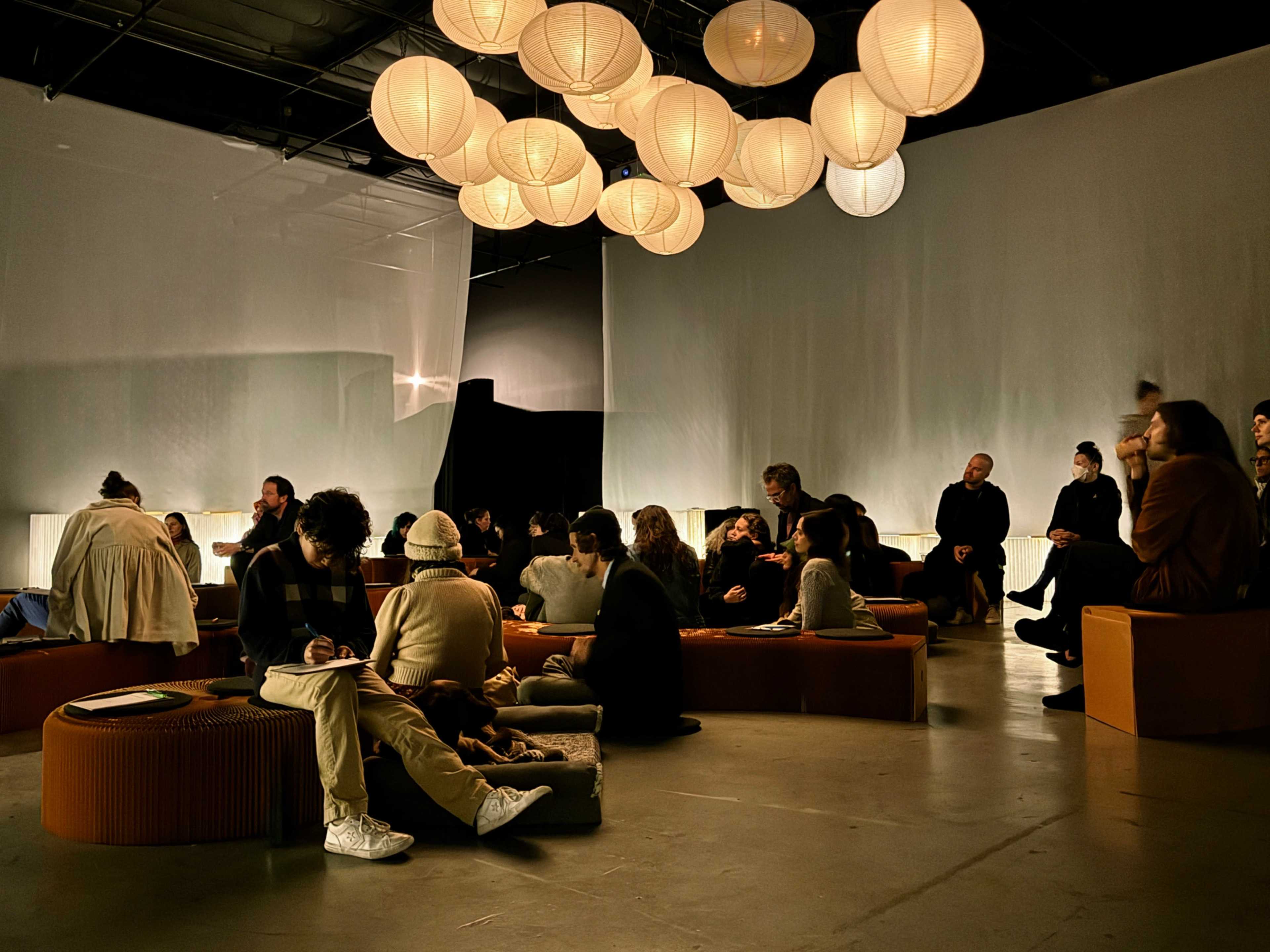 The image depicts a spacious indoor setting with various groups of people seated on benches, illuminated by round paper lanterns hanging from the ceiling.