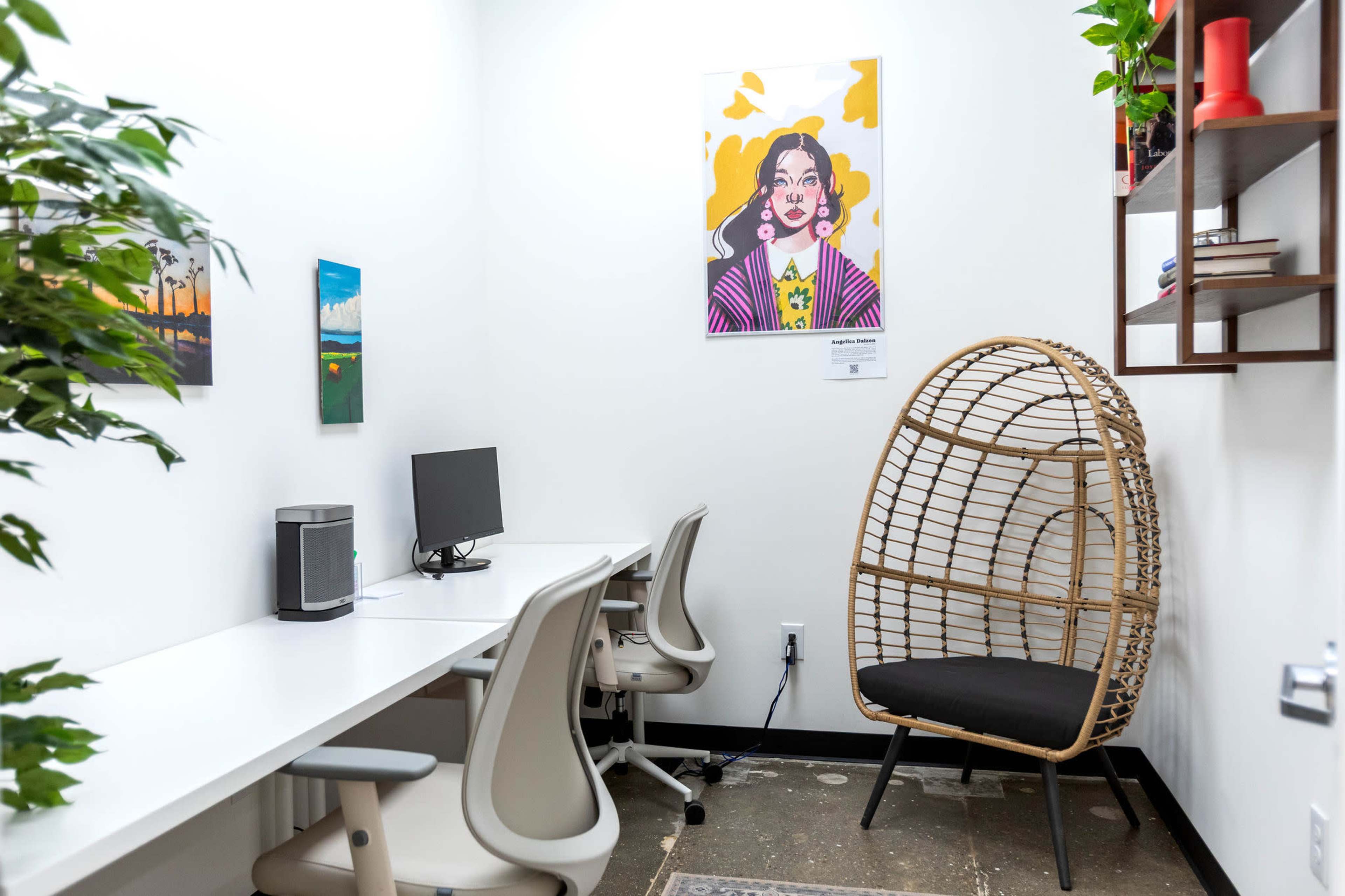 The image shows a small office space with two white desks and chairs, a computer monitor, a decorative plant, and a rattan chair beside a framed artwork on the wall.