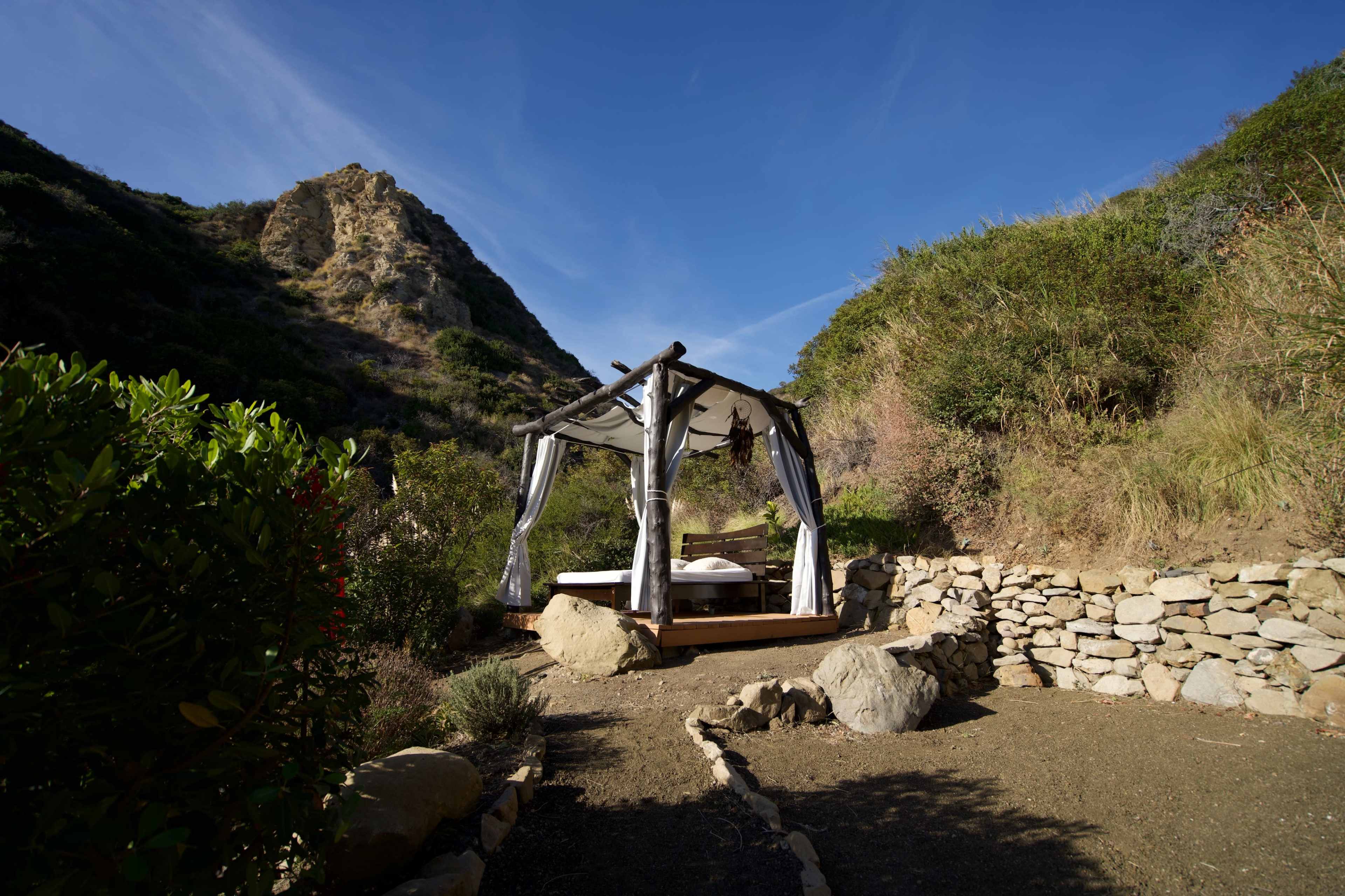 A wooden bed with canopy is situated in a rocky clearing surrounded by bushes, with a mountainous background under a clear blue sky.