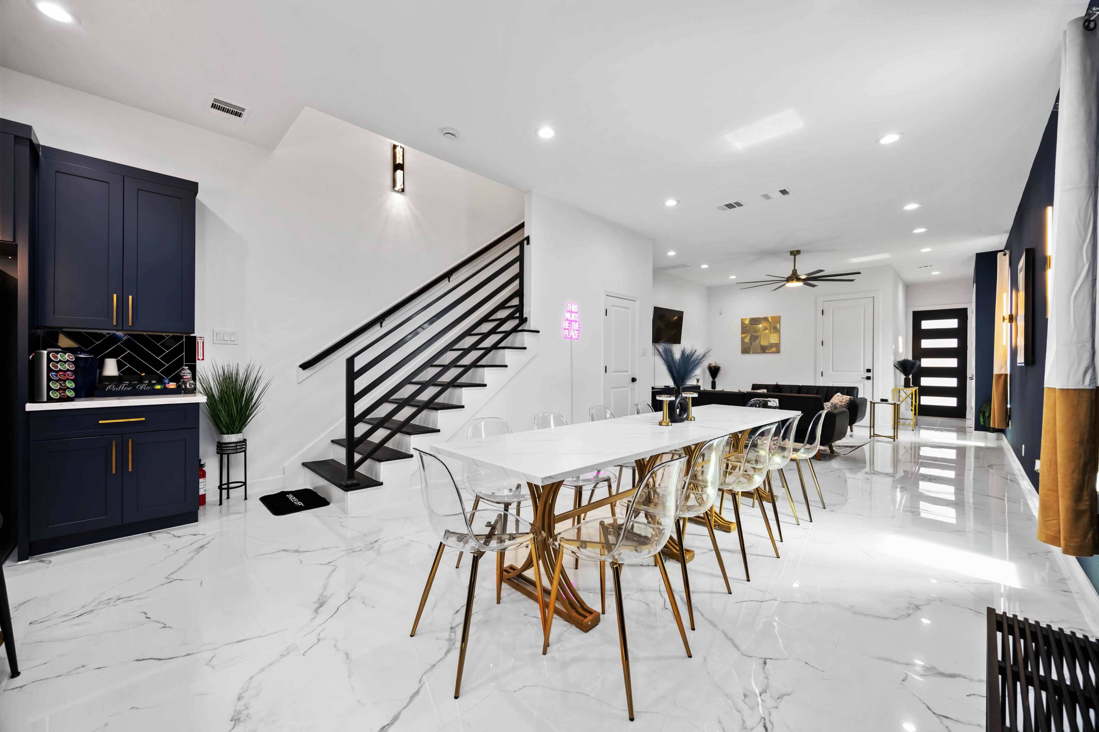 The image shows a modern open-concept kitchen and dining area featuring a white marble floor, a long dining table with transparent chairs, and a staircase leading to an upper level.