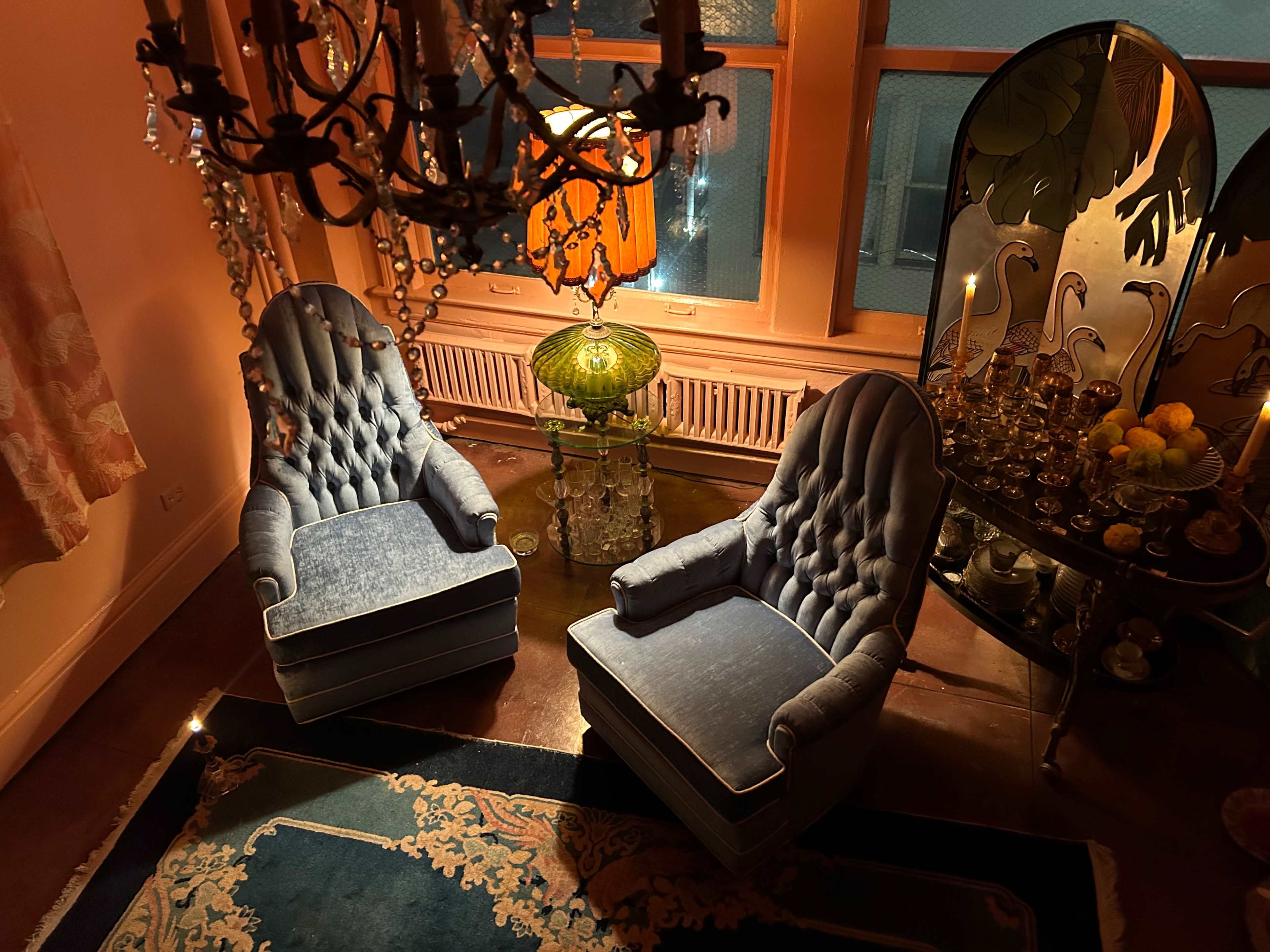 The image shows two upholstered blue chairs facing each other in a warmly lit room with a chandelier and decorative glass table.