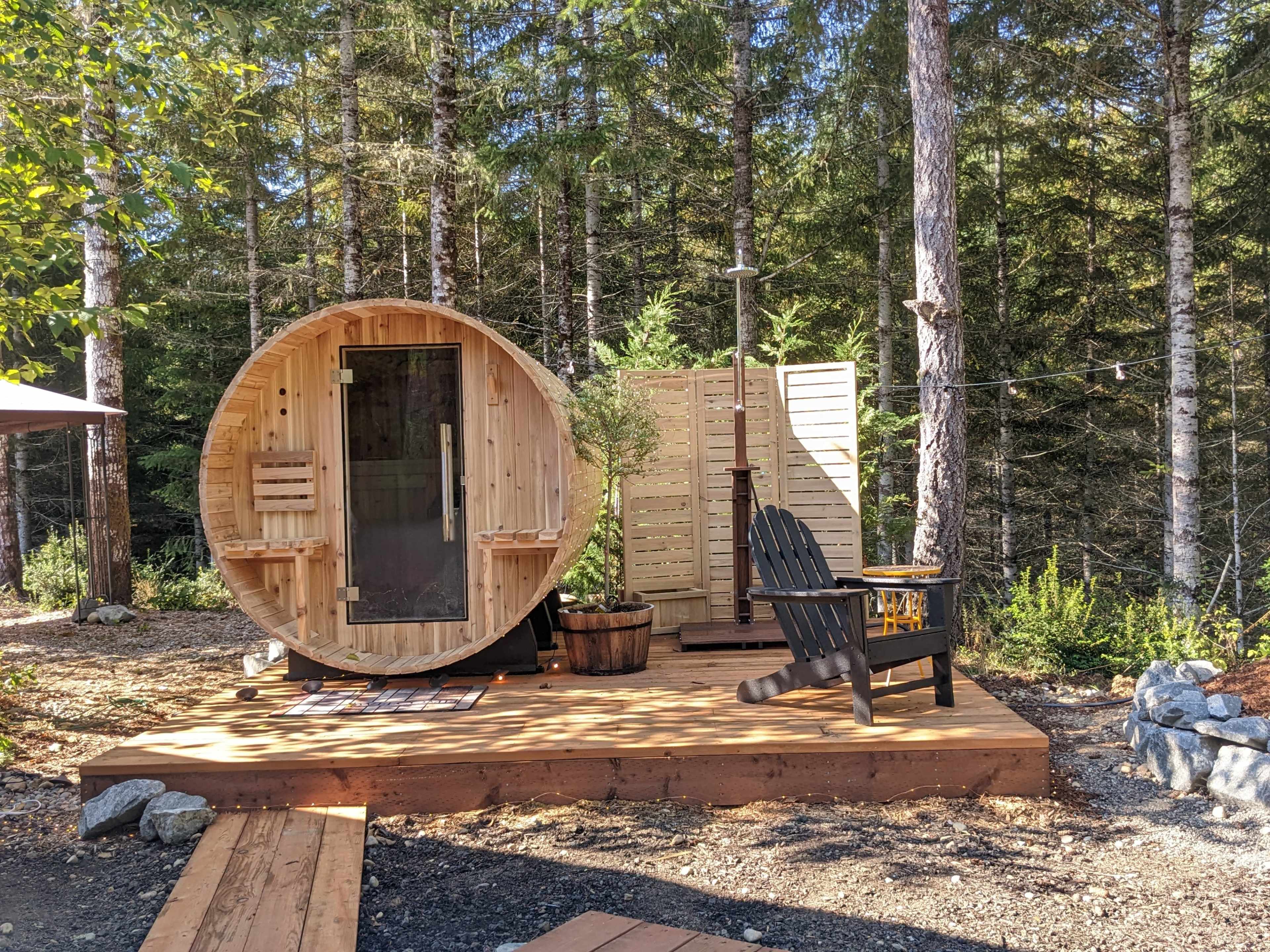 A circular wooden sauna is set on a deck surrounded by trees, with seating and a privacy screen nearby.
