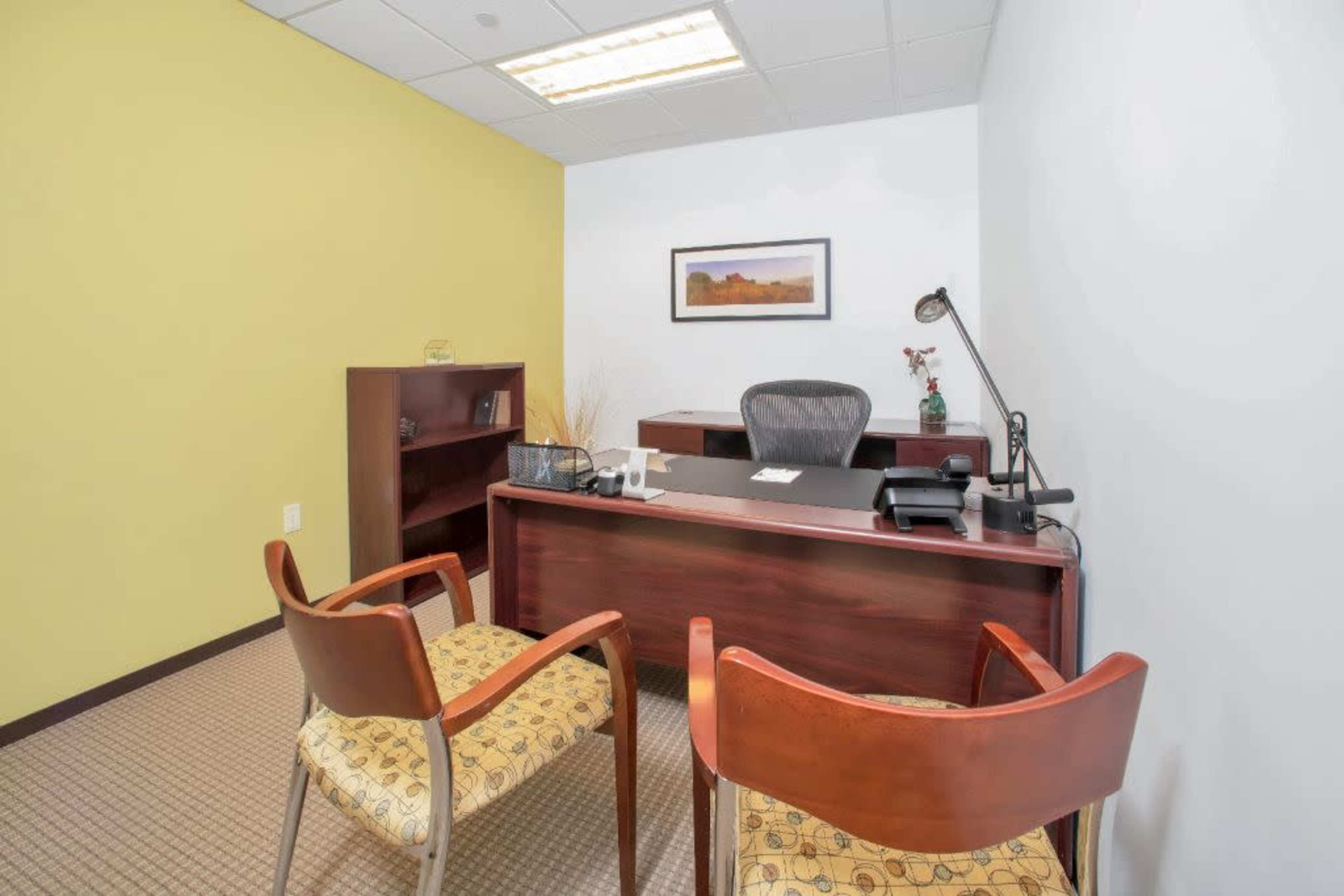 A small office space featuring a desk with a chair, a bookshelf, and two chairs in front of the desk, with a light yellow wall in the background.