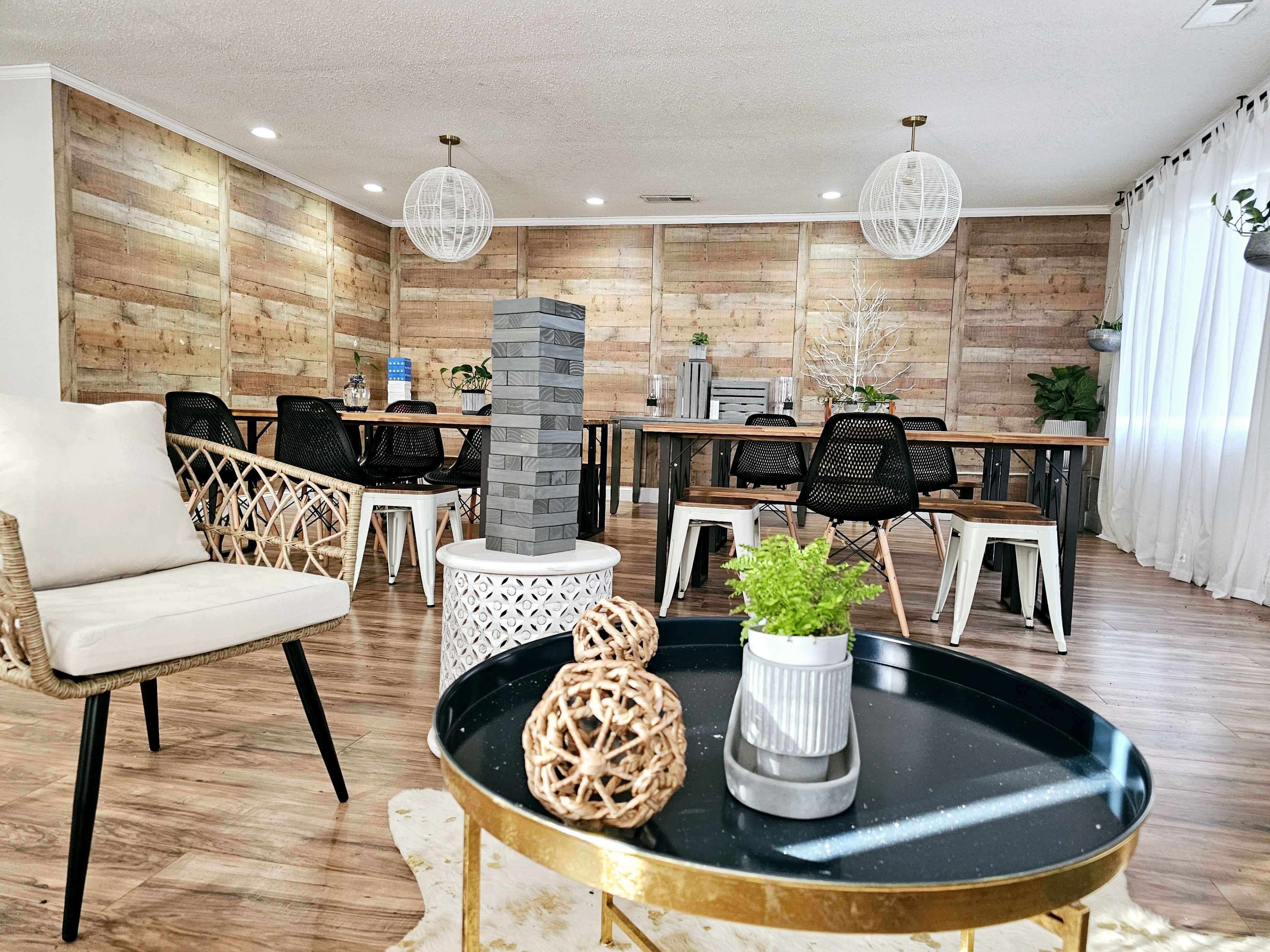 A spacious gathering room with wooden panel walls, a large dining table surrounded by chairs, and decorative elements like plants and a game tower in the foreground.