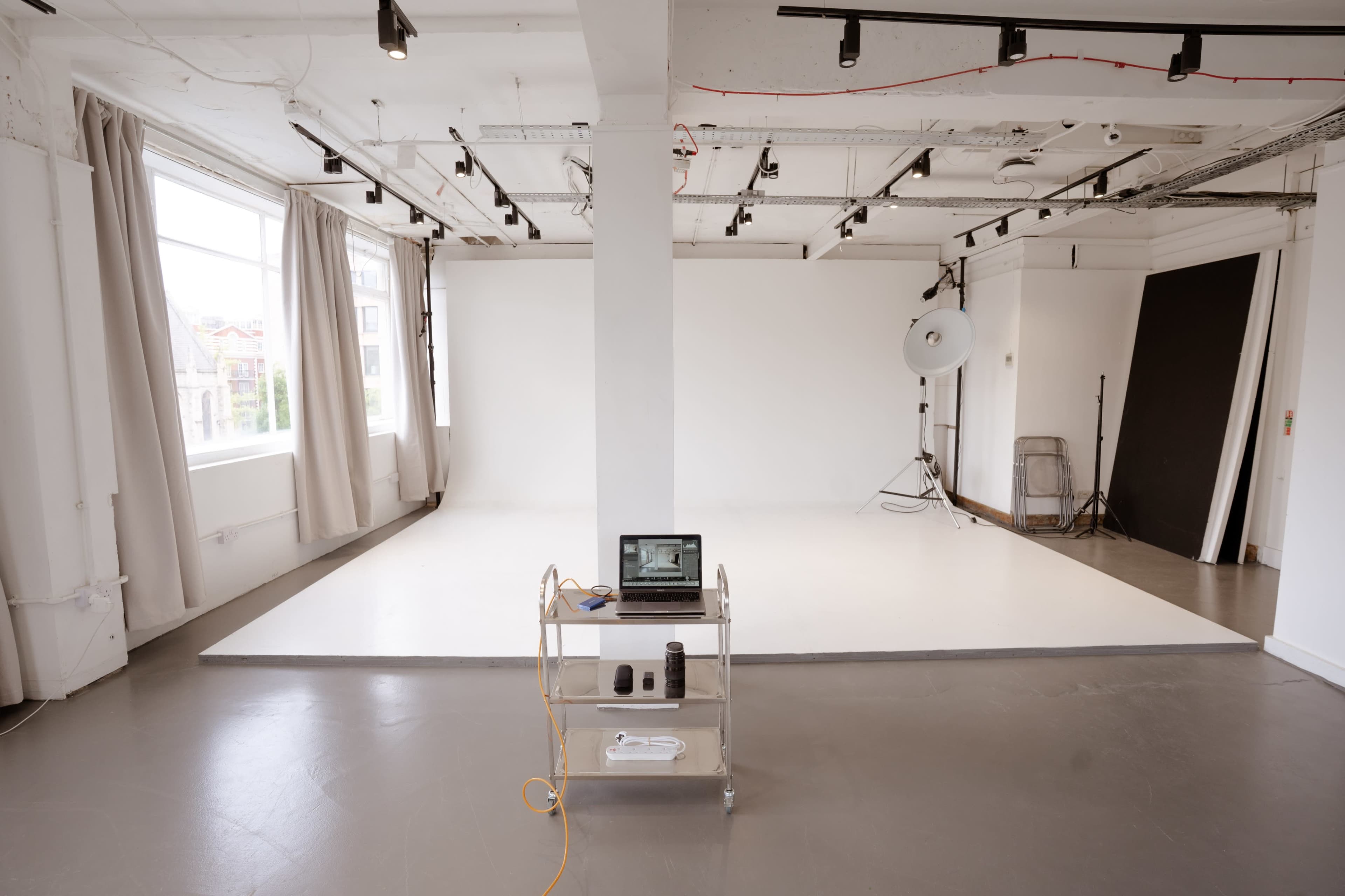 The image shows a spacious, well-lit photography studio featuring a plain white backdrop, a laptop on a cart, and studio lighting equipment.