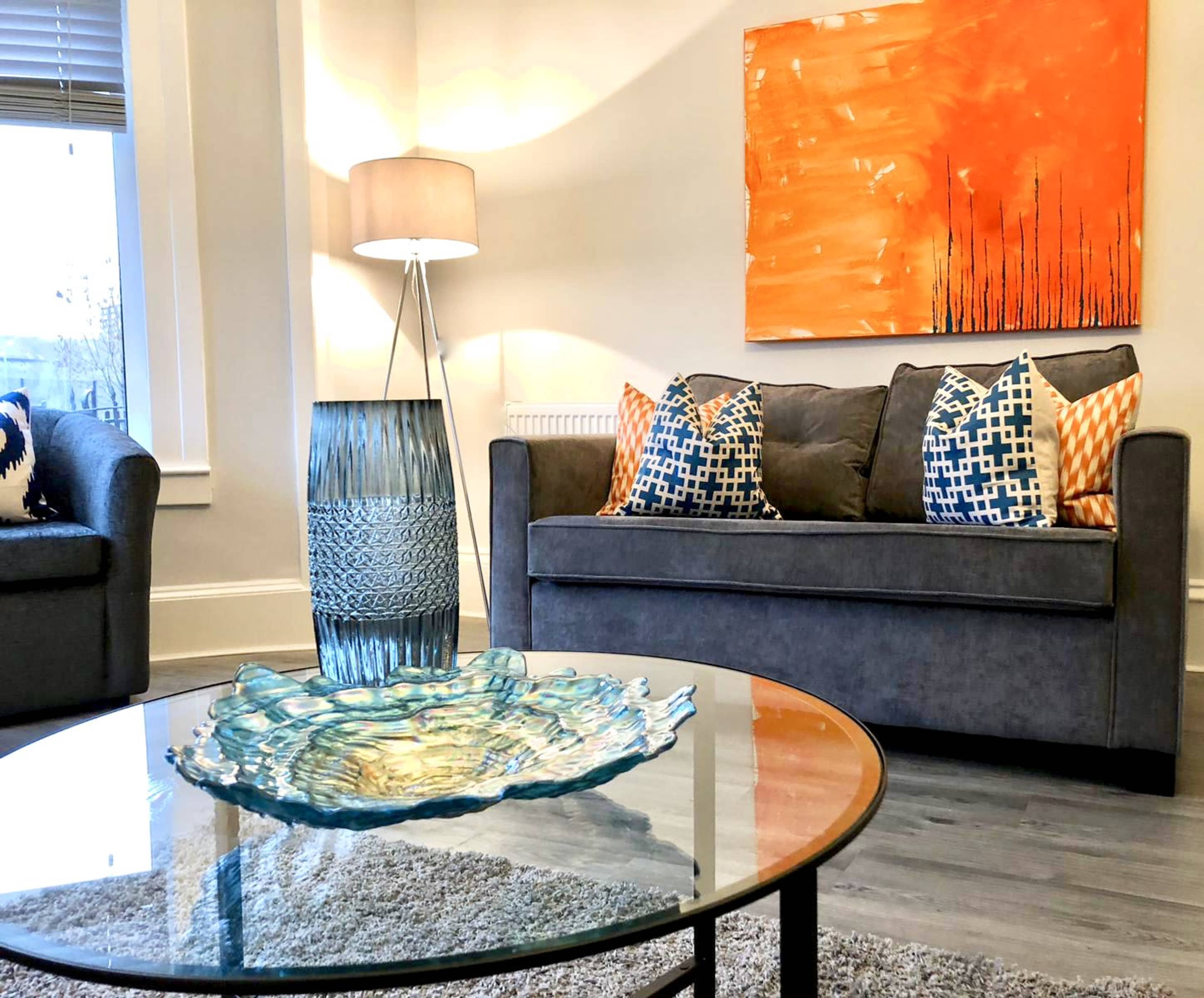 A living room features a gray sofa with decorative pillows, an orange abstract painting on the wall, a glass coffee table, and a blue decorative vase.
