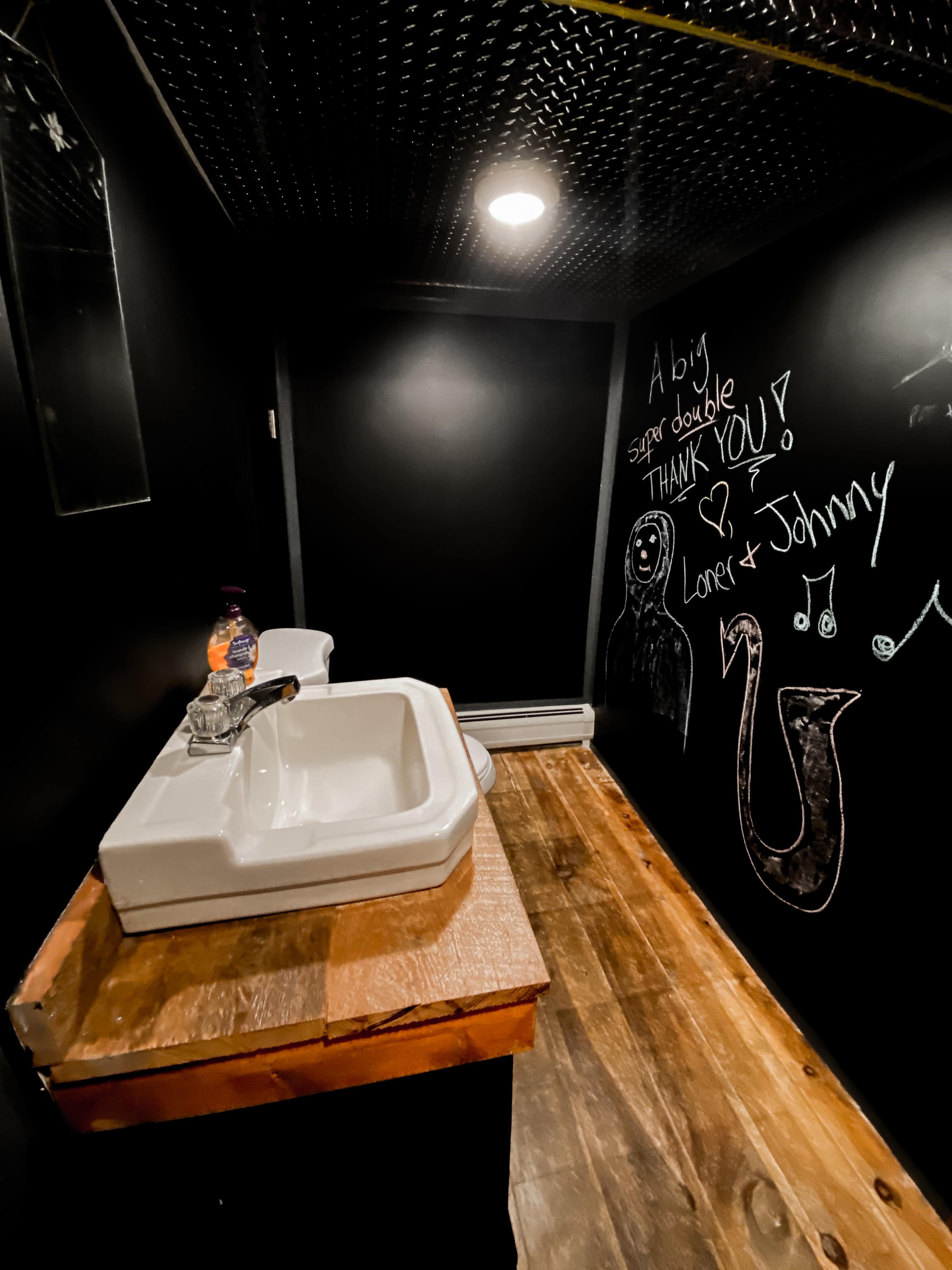 The image shows a small, dark bathroom with a white sink on a wooden countertop and a wall covered in chalkboard paint featuring handwritten messages and drawings.