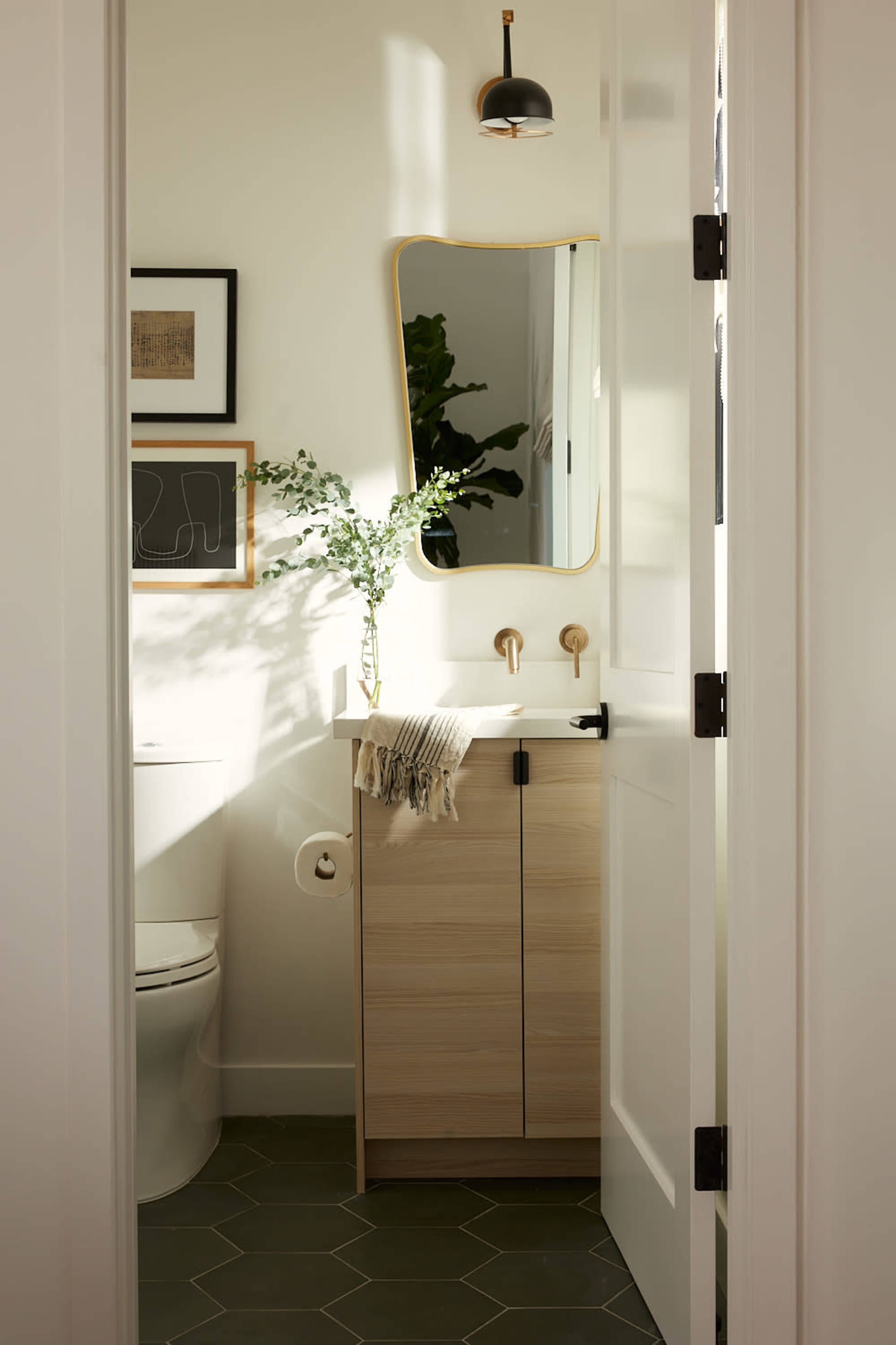 A well-lit bathroom features a light wood cabinet, a round mirror, and a potted plant beside the toilet.