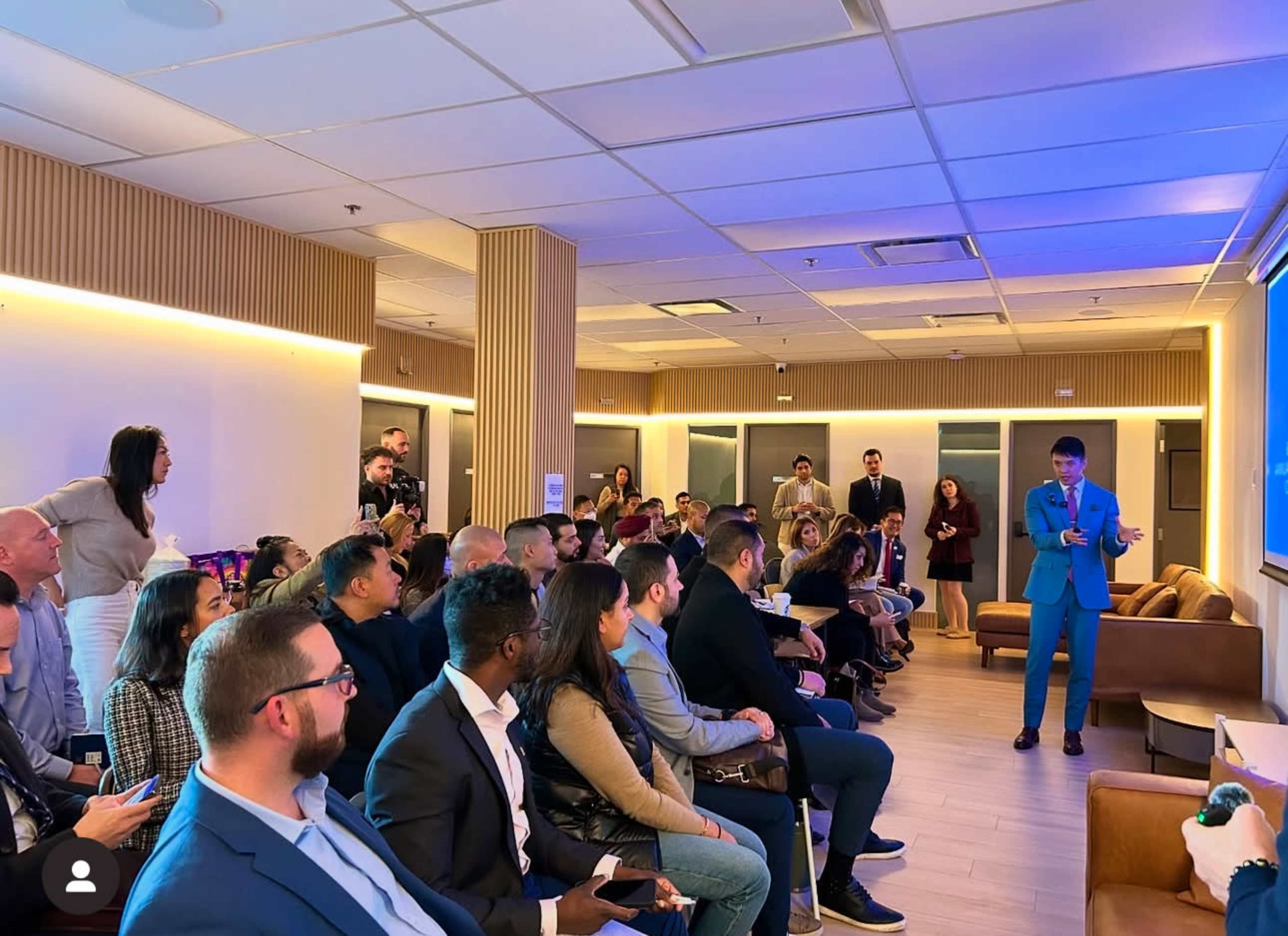 A speaker in a blue suit presents in front of a seated audience in a well-lit, modern conference room.