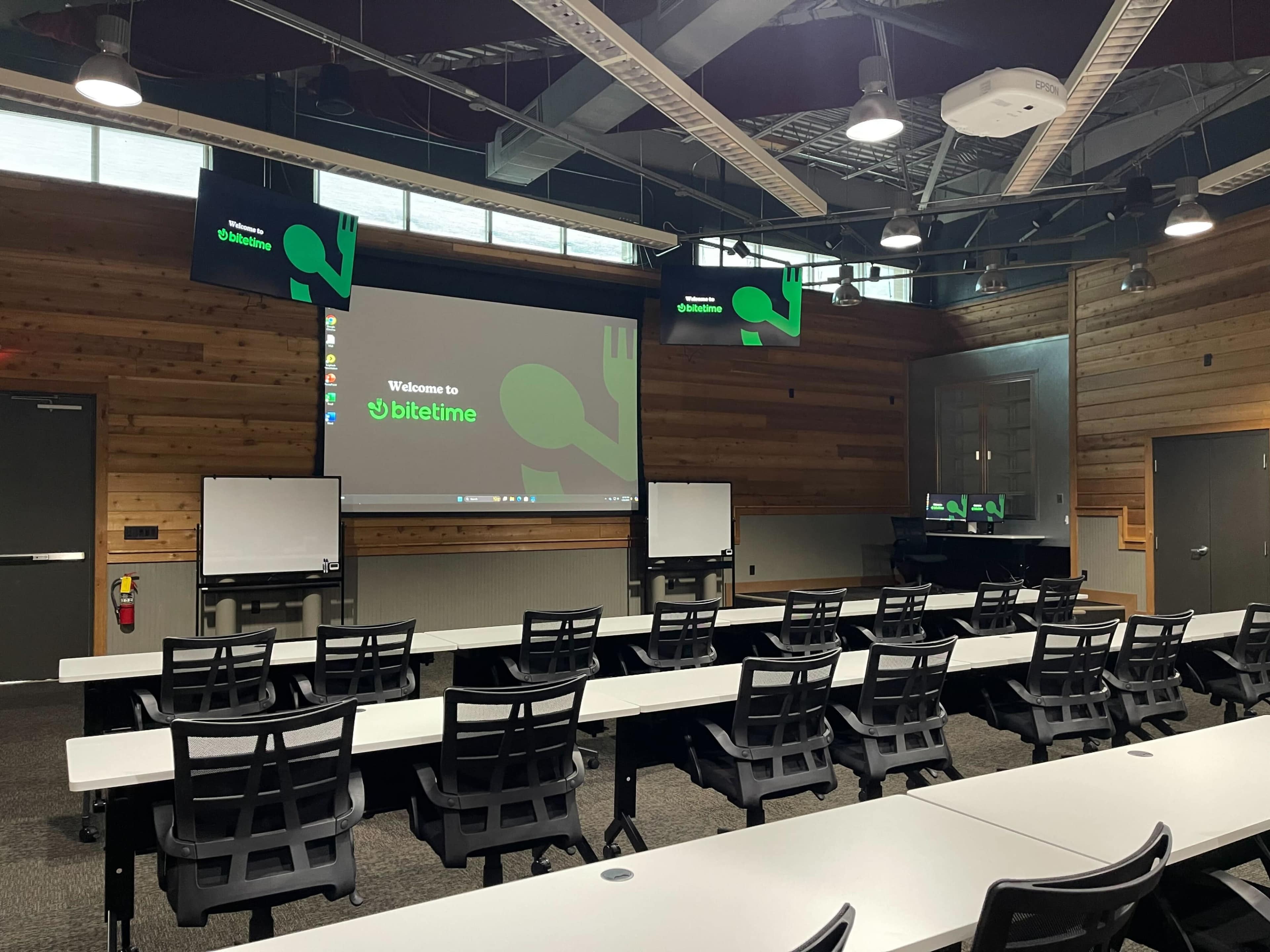 The image shows a modern classroom or conference room with rows of black chairs and white tables, equipped with a large screen displaying a welcome message and multiple monitors on the walls.