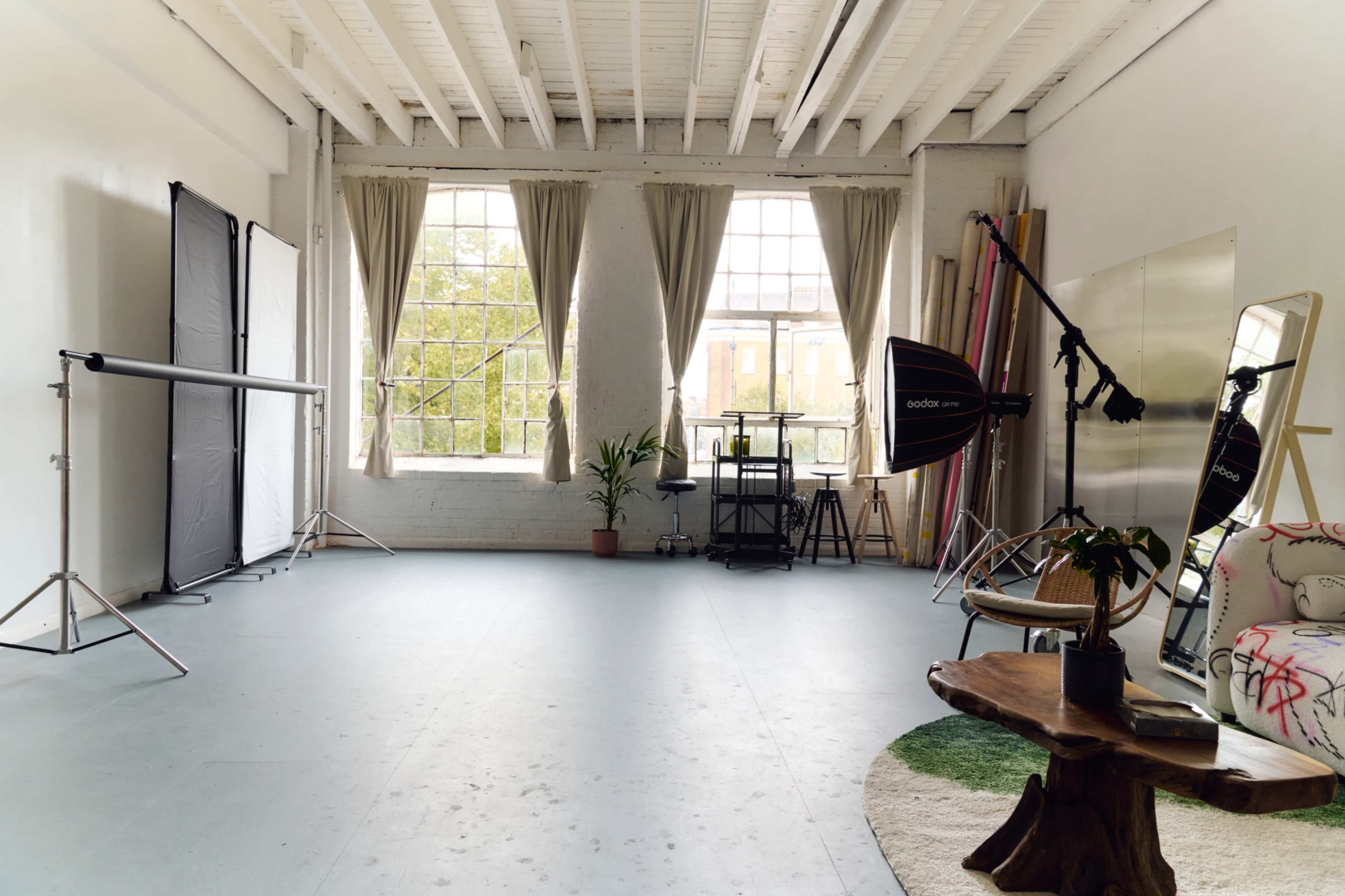 The image depicts a spacious photography studio featuring large windows, equipment like light stands and a reflector, a potted plant, and a few pieces of furniture.