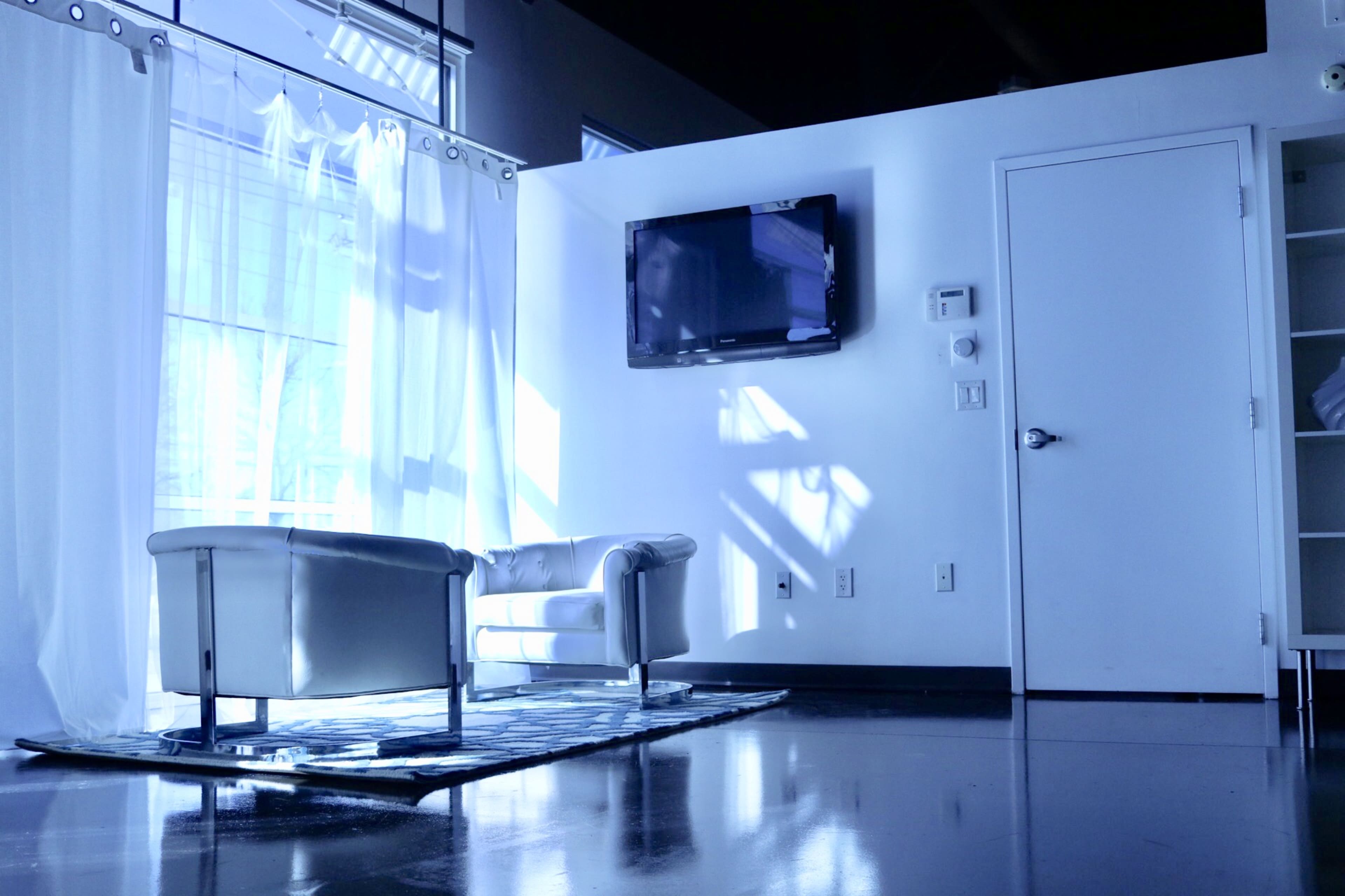 A light-filled room features a white couch, a television mounted on the wall, and a door leading to another space.