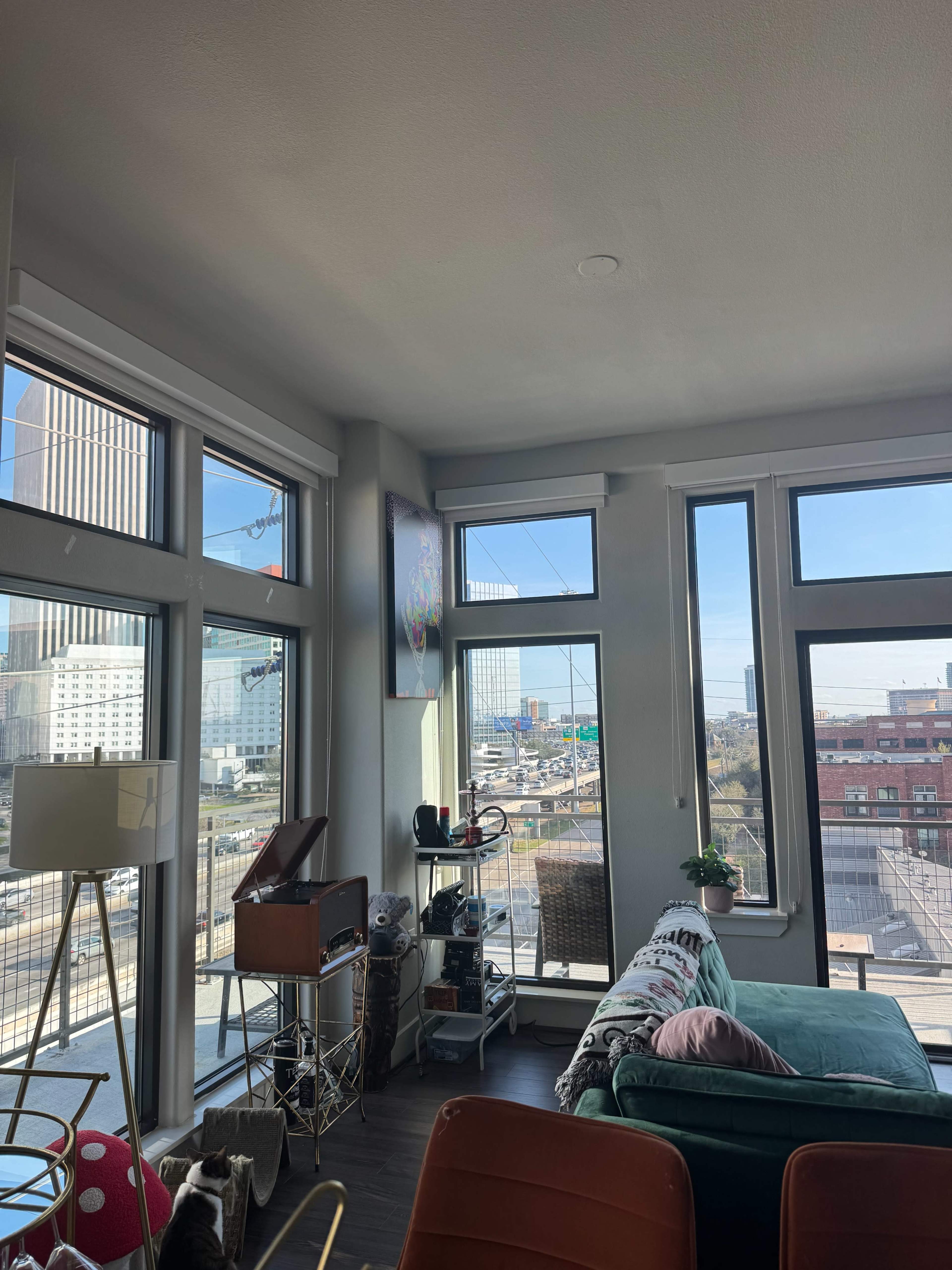 The image shows a modern living room with large windows overlooking a cityscape, featuring a couch, a record player, and decorative plants.