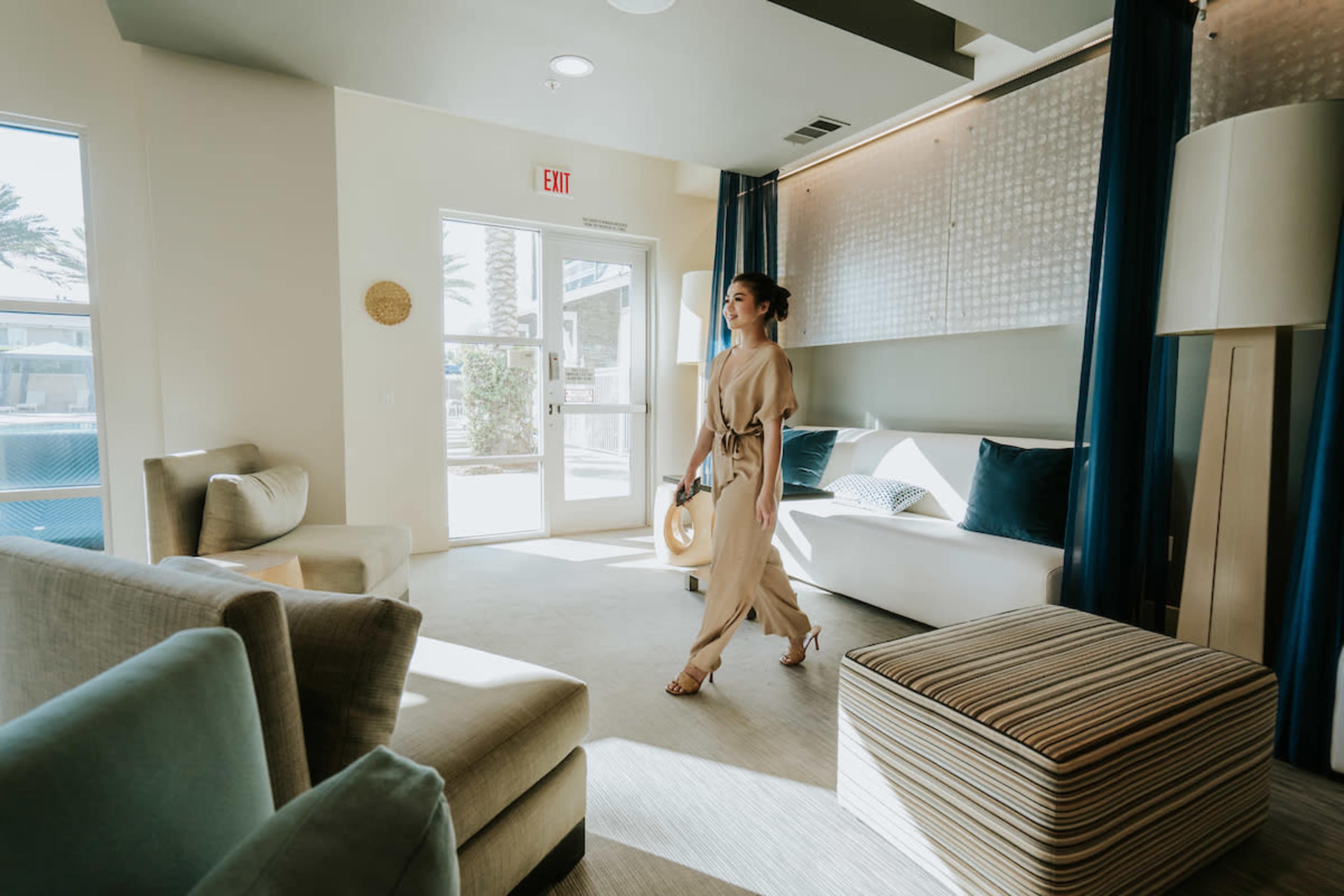 A woman walks through a modern lounge area with light-colored furniture and large windows.