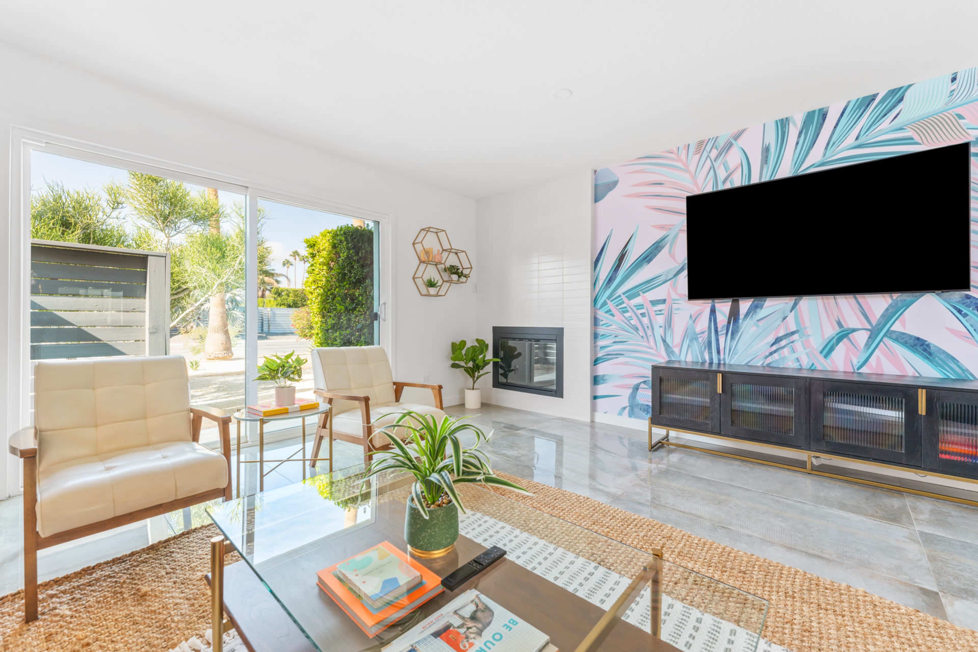 A modern living room with two chairs, a coffee table, and a wall featuring tropical-themed wallpaper, along with a fireplace and a large window leading to an outdoor view.