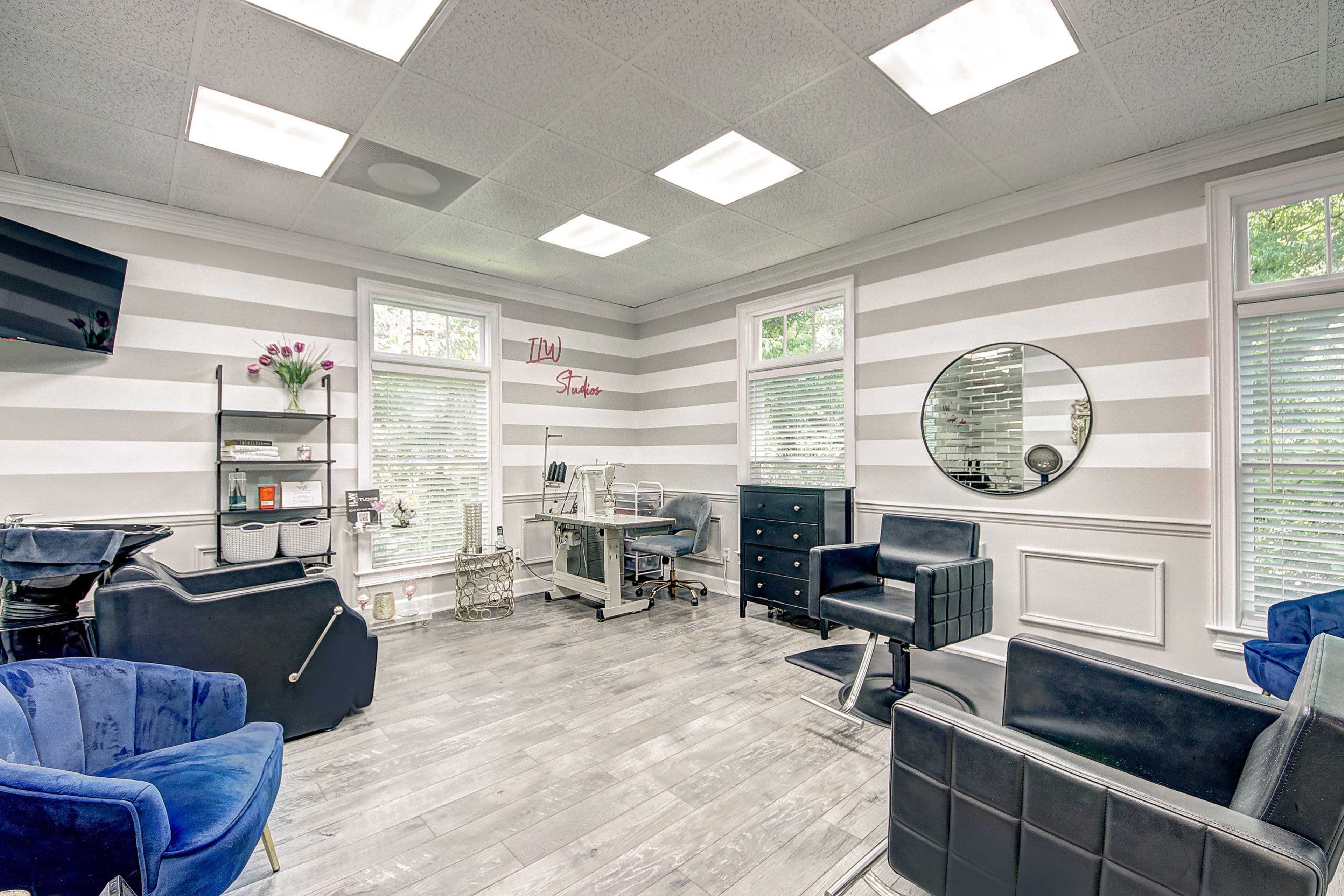 A modern salon interior with striped walls, a central hair styling station, and black and blue chairs arranged around the space.