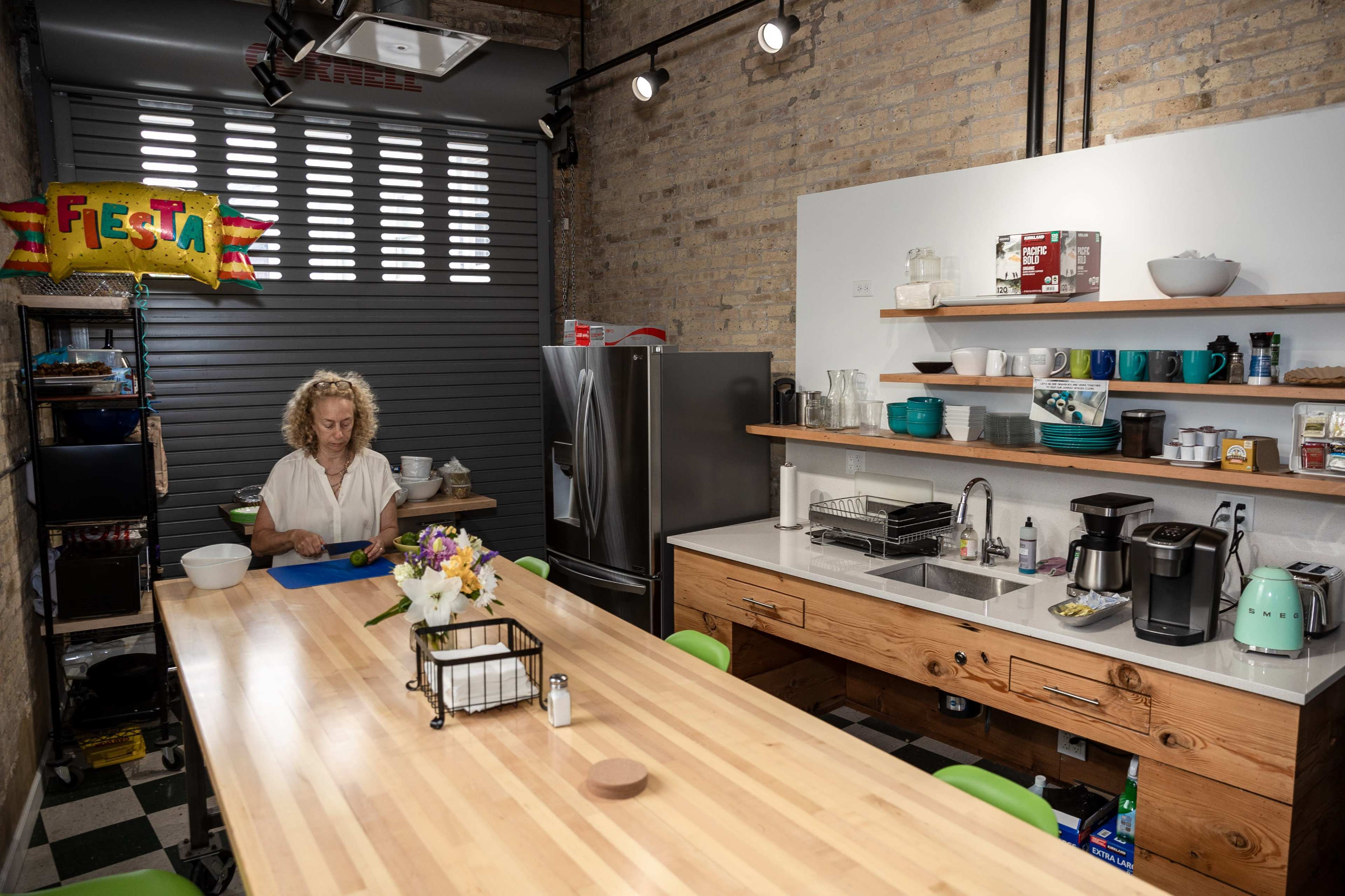 A woman prepares food at a wooden table in a modern kitchen with various kitchen appliances and colorful decorations.