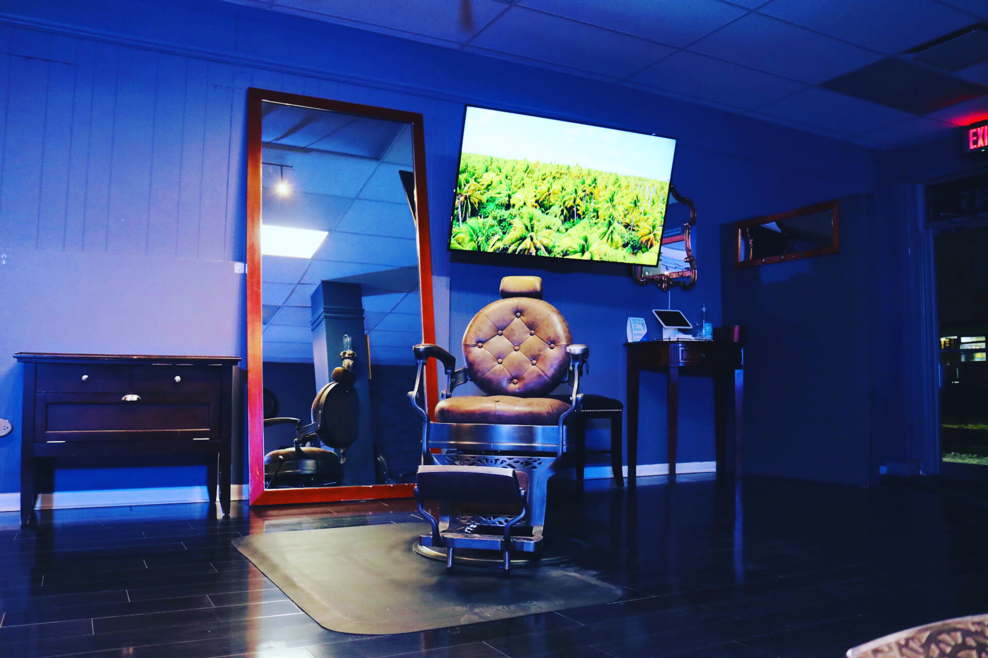 A barbershop interior featuring a leather barber chair, a large mirror, a television displaying a landscape, and a dark wooden cabinet in a blue-lit room.