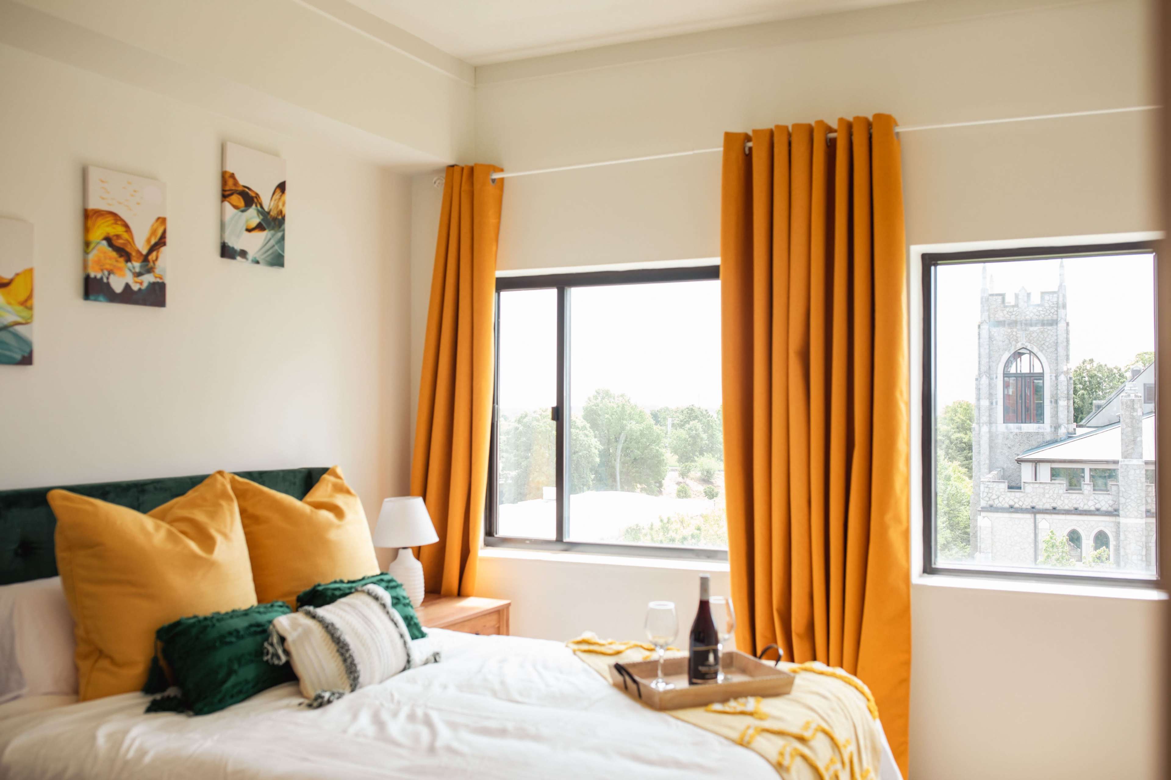 A bright bedroom features a bed with yellow and green pillows, large windows with orange curtains, and views of a stone tower outside.