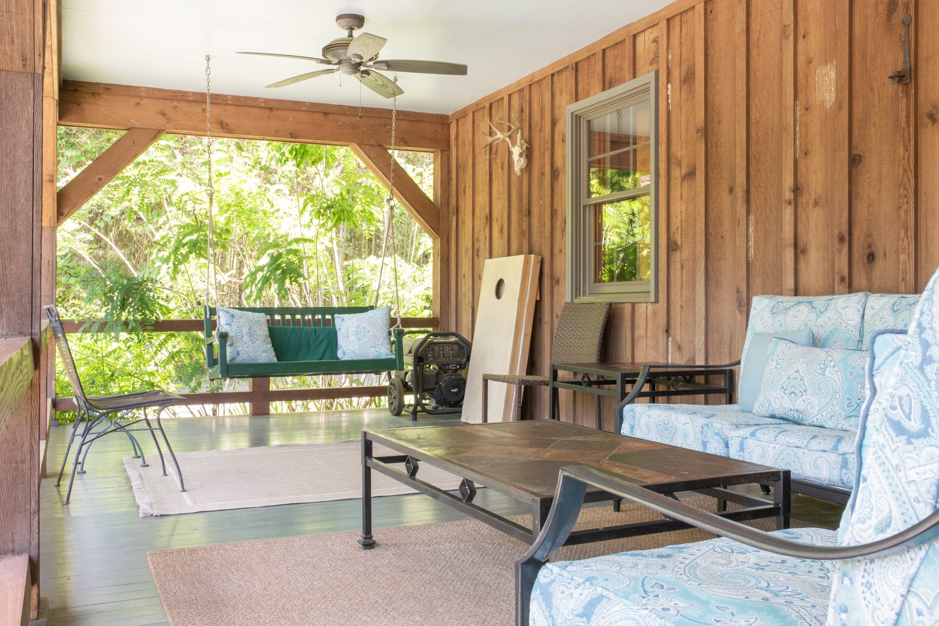 The image shows a porch with wooden walls, a ceiling fan, a swing, and various seating arrangements around a central coffee table.