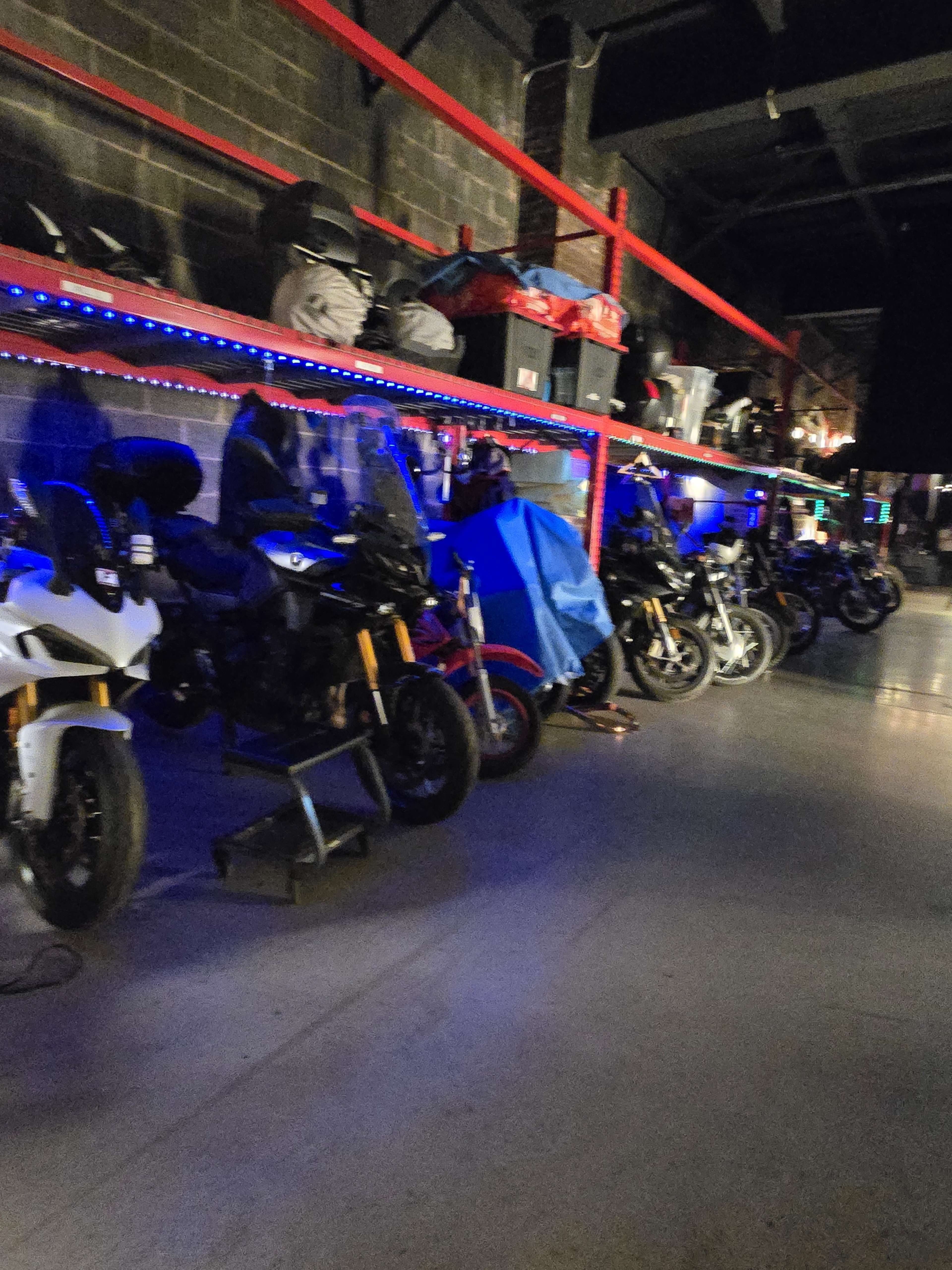 The image shows a row of motorcycles parked under a shelf illuminated with blue lights in a dimly lit space.