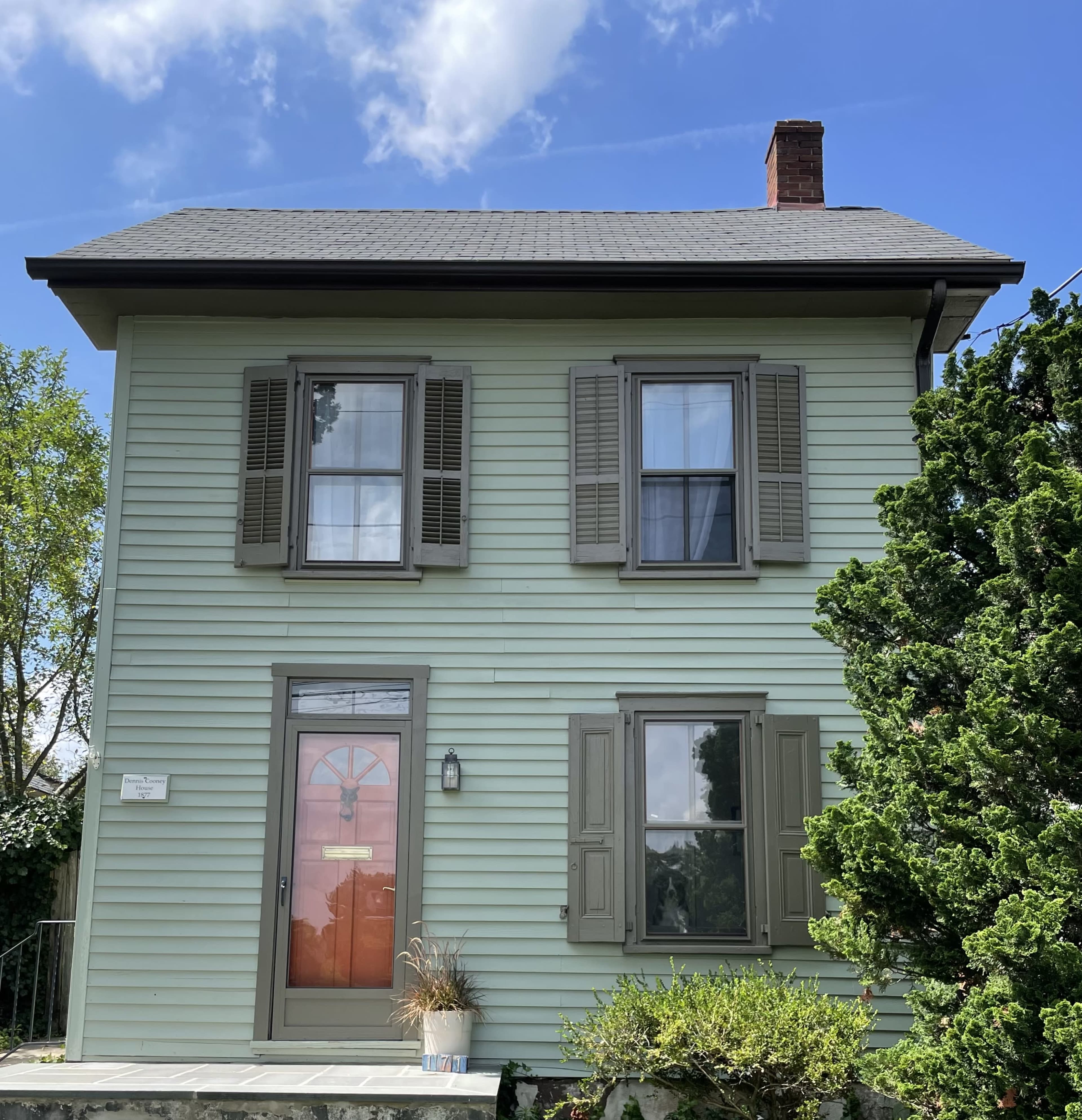 A two-story, pale green house with a front door flanked by two windows and surrounded by shrubs and a small tree.