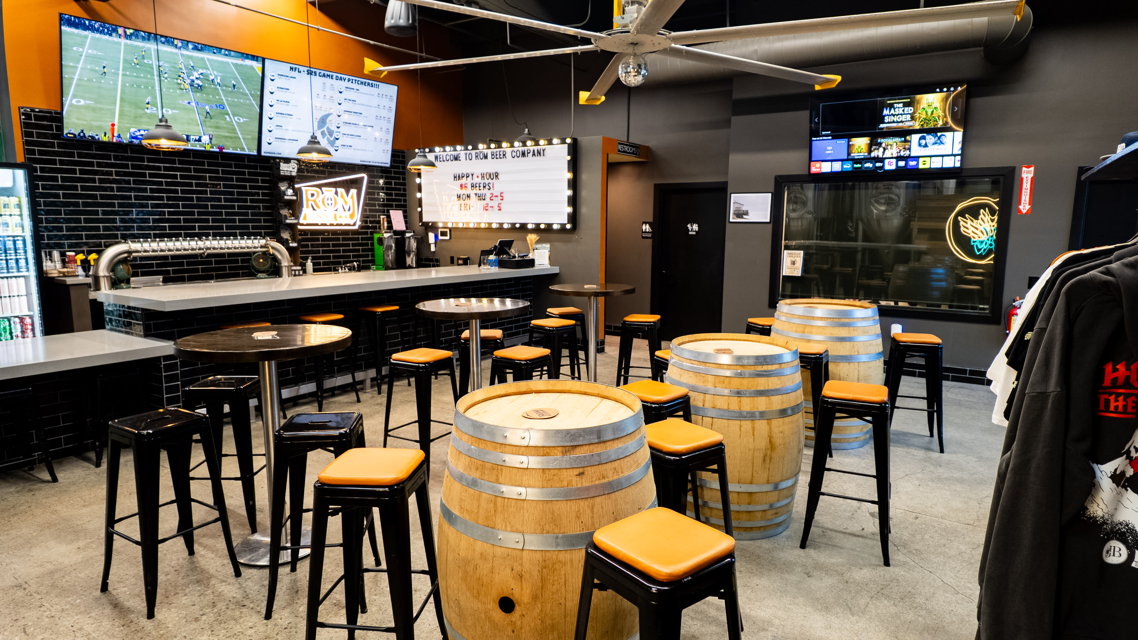 The image shows a modern bar interior with wooden barrel tables, high stools, and multiple screens displaying sports events.