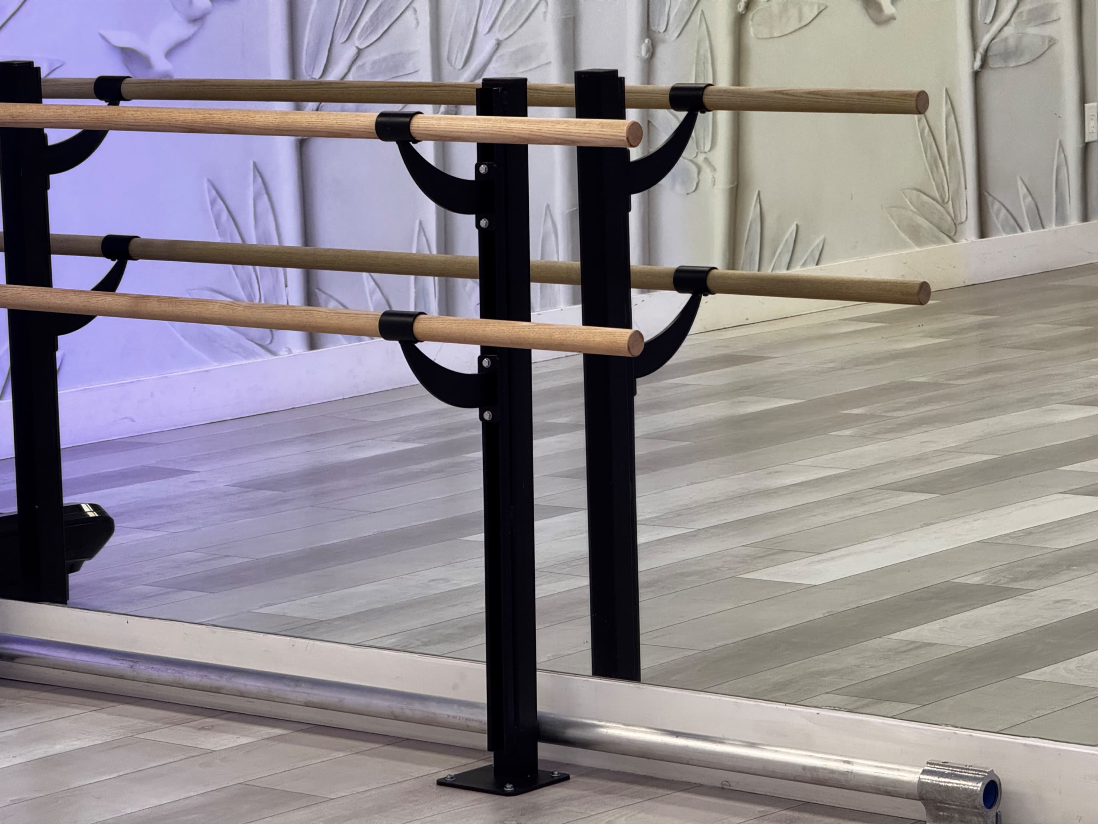 The image shows a ballet barre made of wood, mounted on black metal supports, positioned against a textured wall in a dance studio.