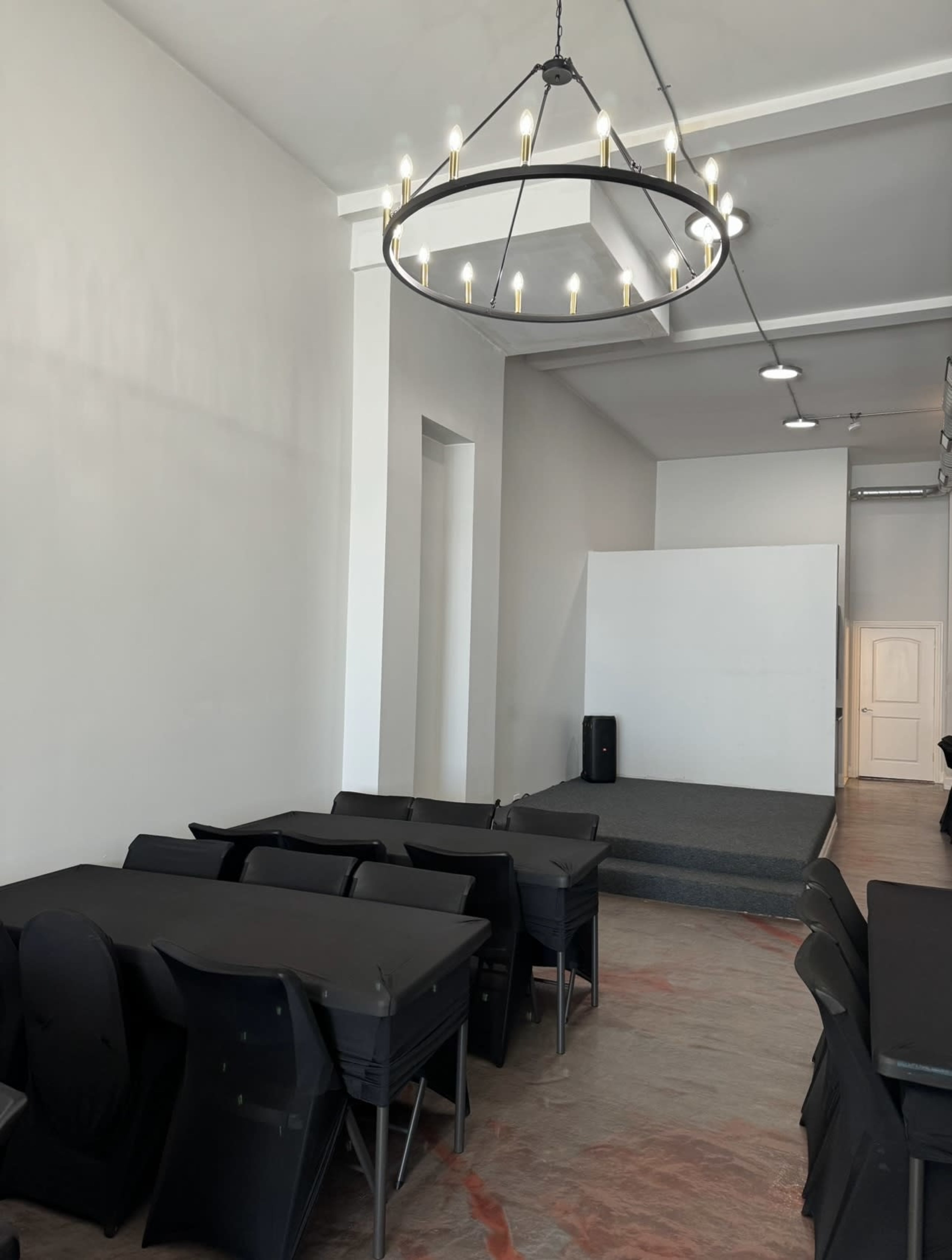 A room with a small stage at the front, several black tables and chairs arranged in the center, and a large circular chandelier hanging from the ceiling.