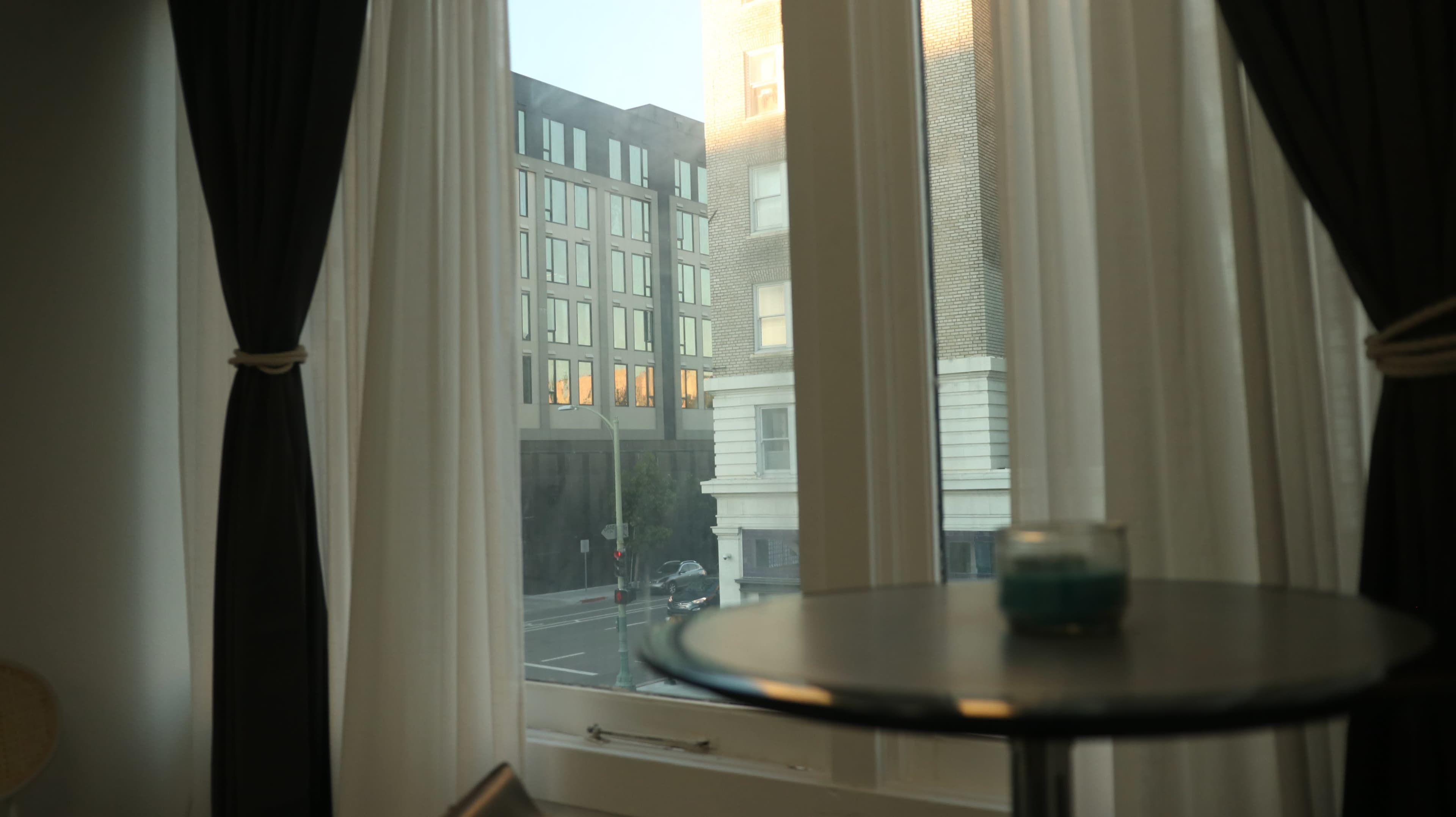 The image shows sheer curtains framing a window that overlooks a city street and a modern building across from the window.