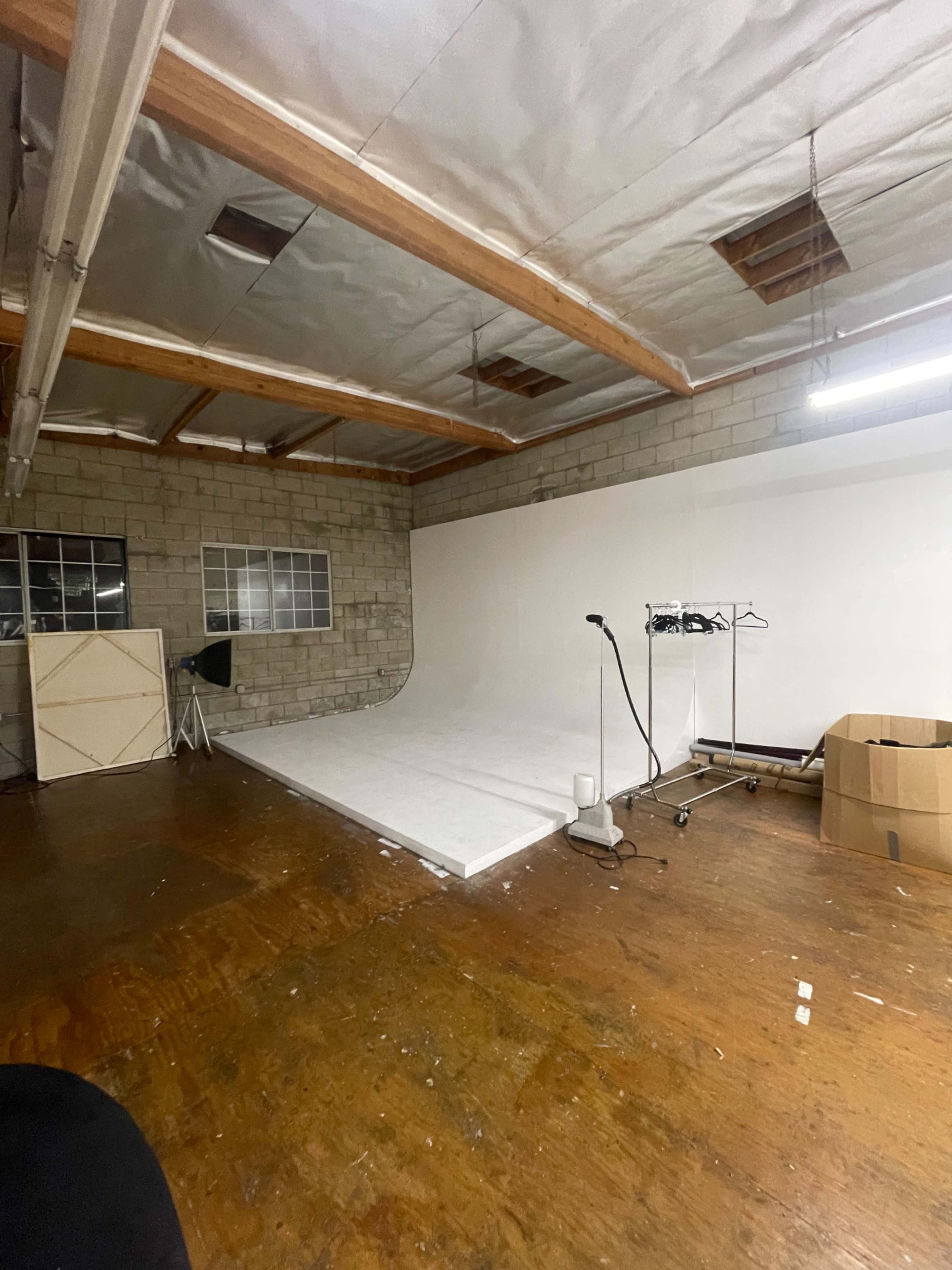 The image shows a photography studio with a white backdrop, a rolling clothing rack, and cardboard boxes in a spacious room with wooden flooring.