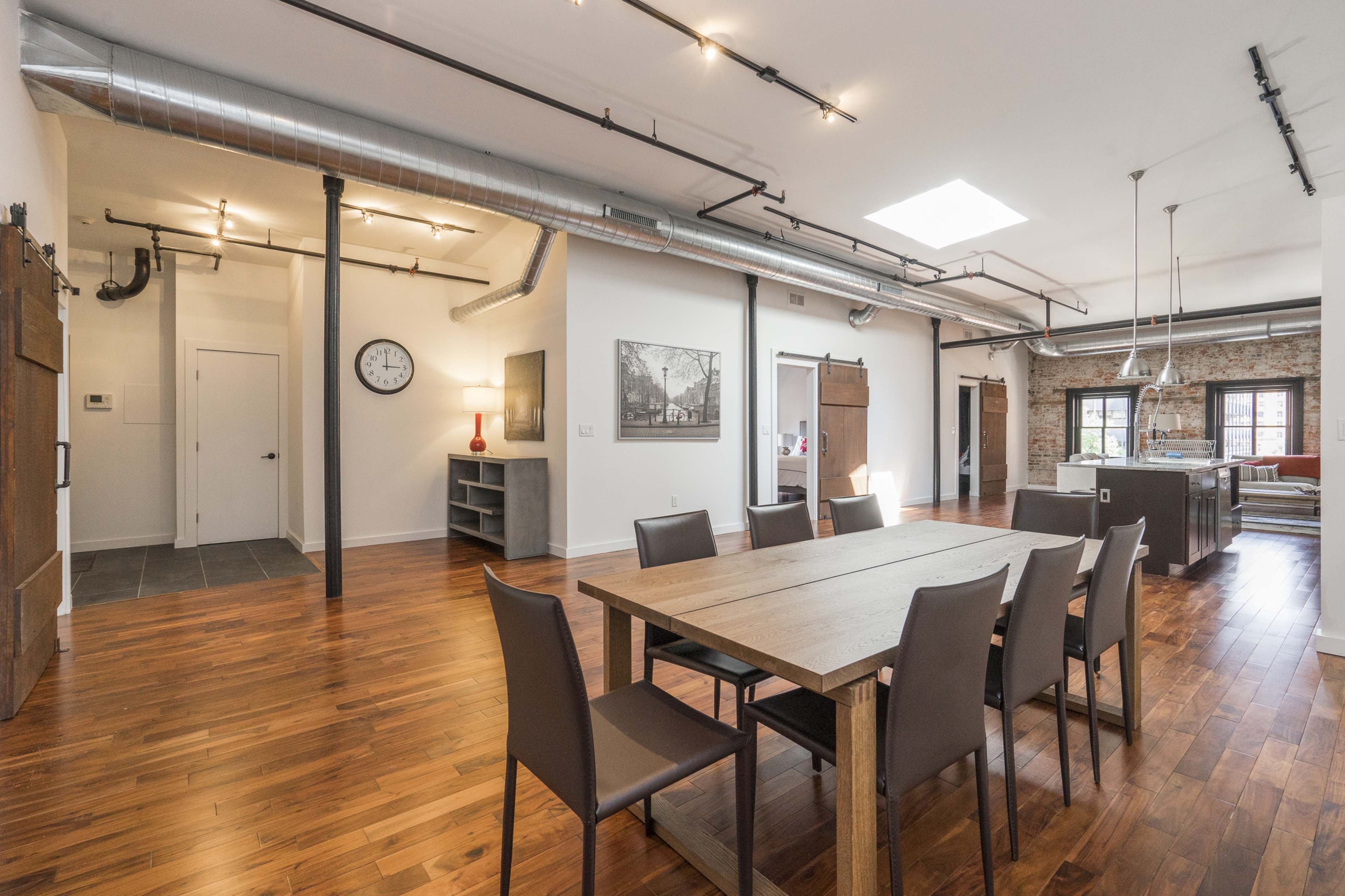 A spacious dining area with a wooden table surrounded by chairs, connected to a modern kitchen and featuring exposed ductwork along the ceiling.