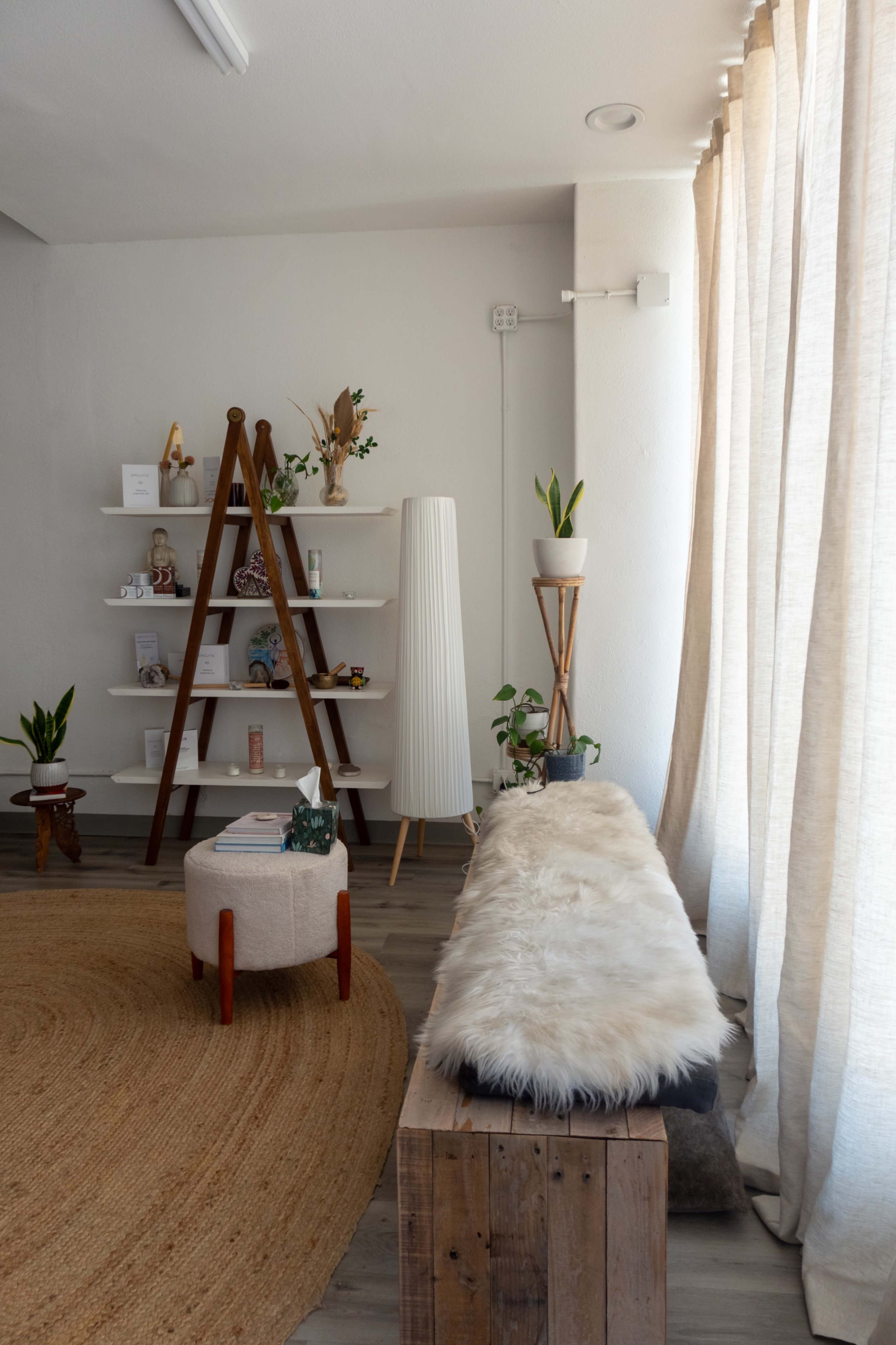 The image shows a cozy interior space featuring a wooden bench with a fur throw, a round rug, a ladder shelf with decorative items, and plants in various pots near large windows with light curtains.