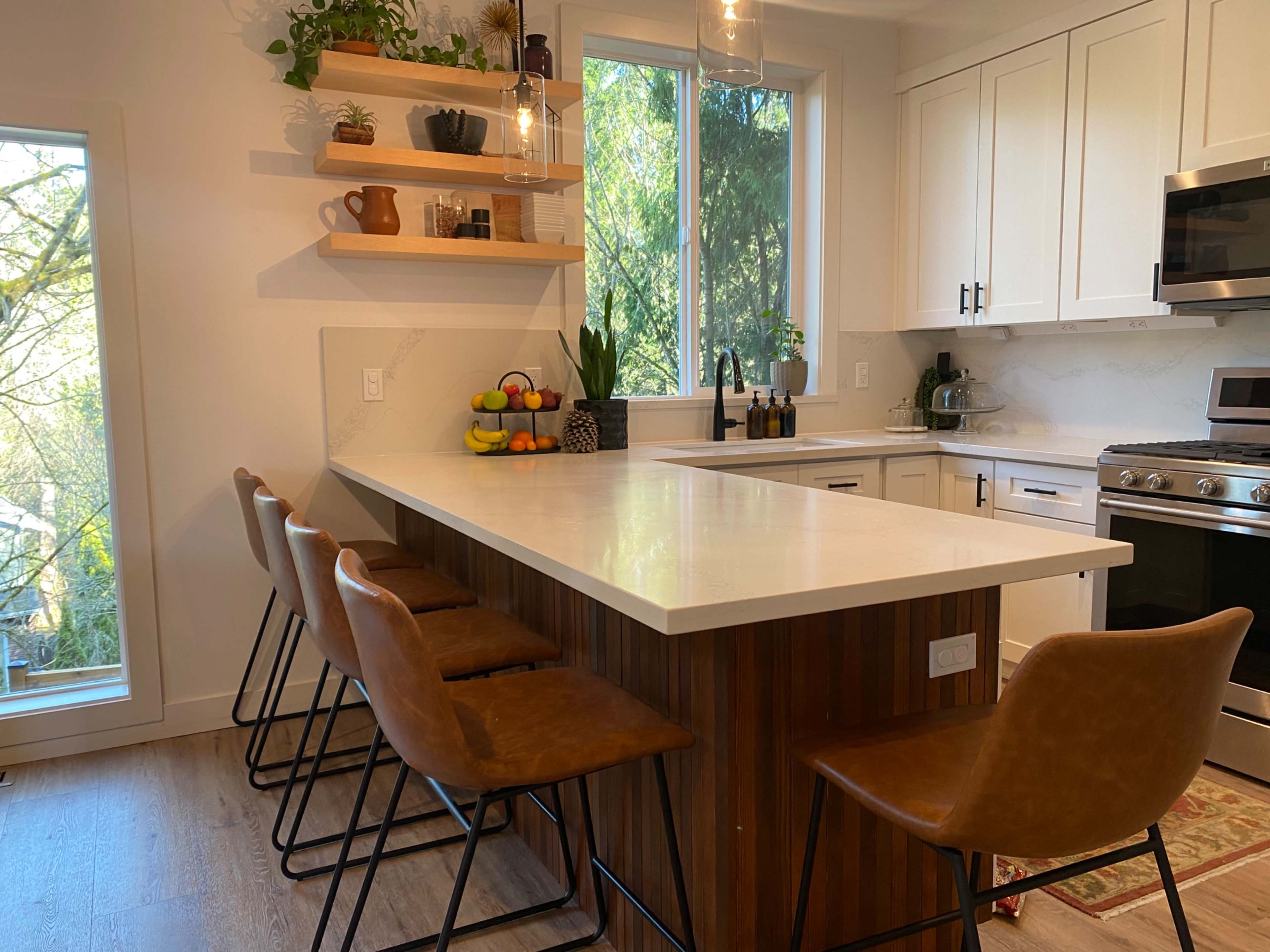 A modern kitchen with a central island featuring bar stools, shelves of plants and decor, and large windows allowing natural light to enter.